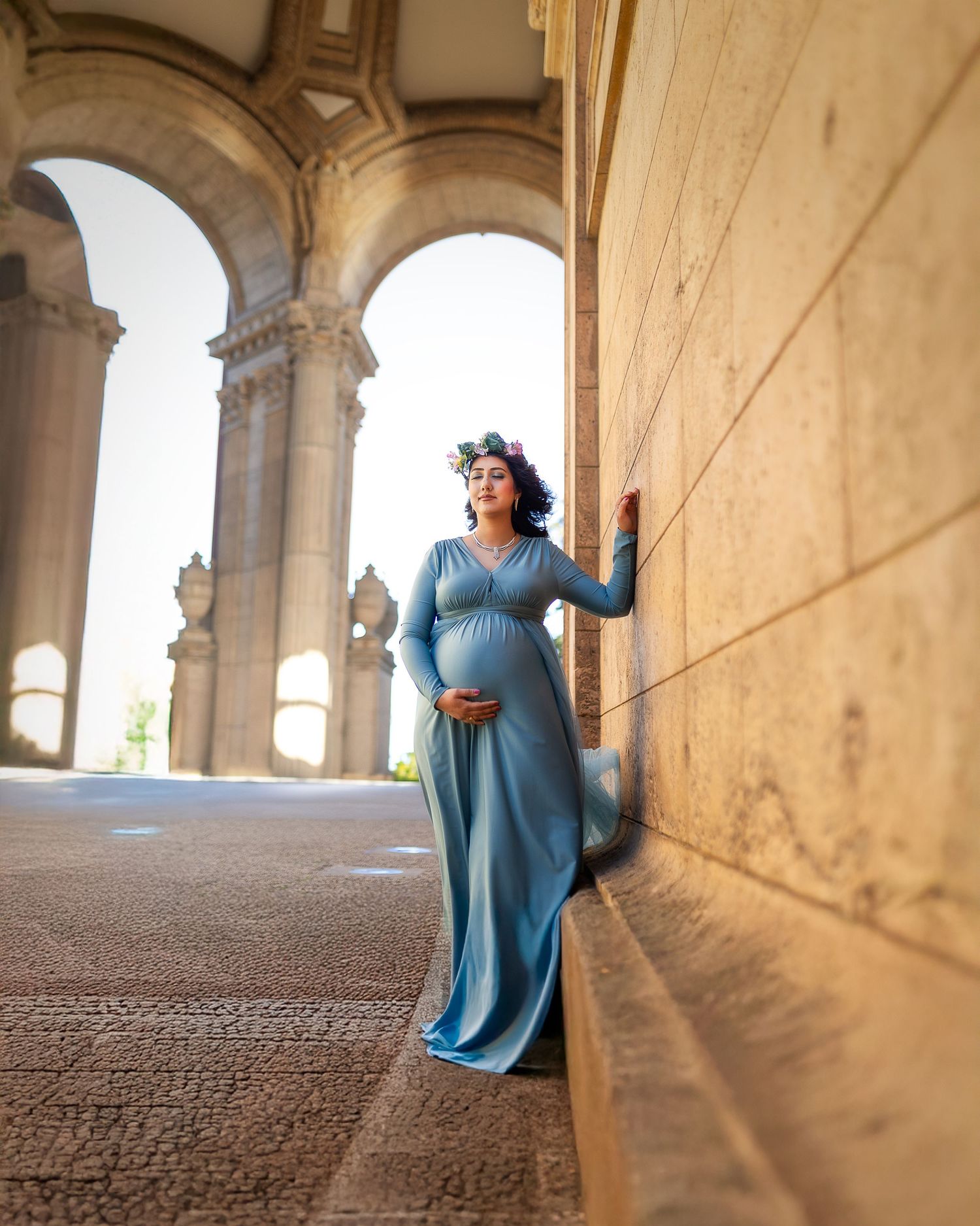 Woman in blue dress poses gracefully under stone archways in dramatic sunlit architectural corridor.