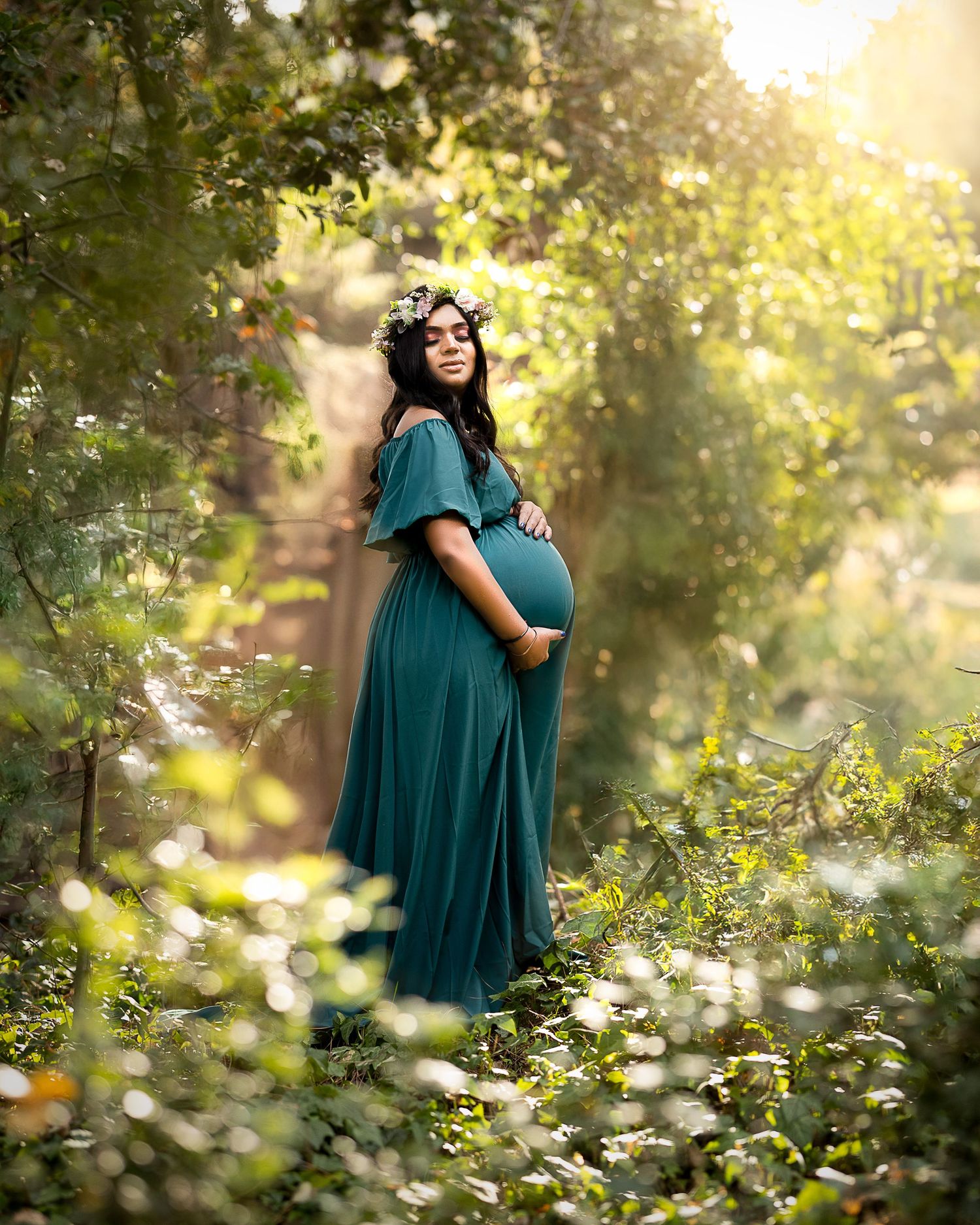 A figure in a flowing teal maternity dress stands among blooming white wildflowers in a sunlit forest setting.