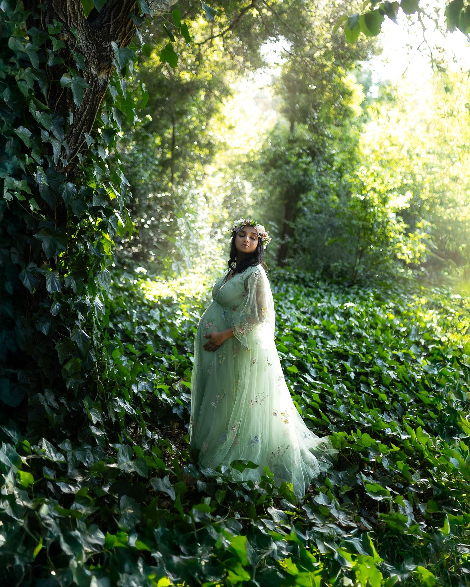 A figure in a flowing mint green dress and veil stands ethereally among lush green foliage in dappled sunlight.