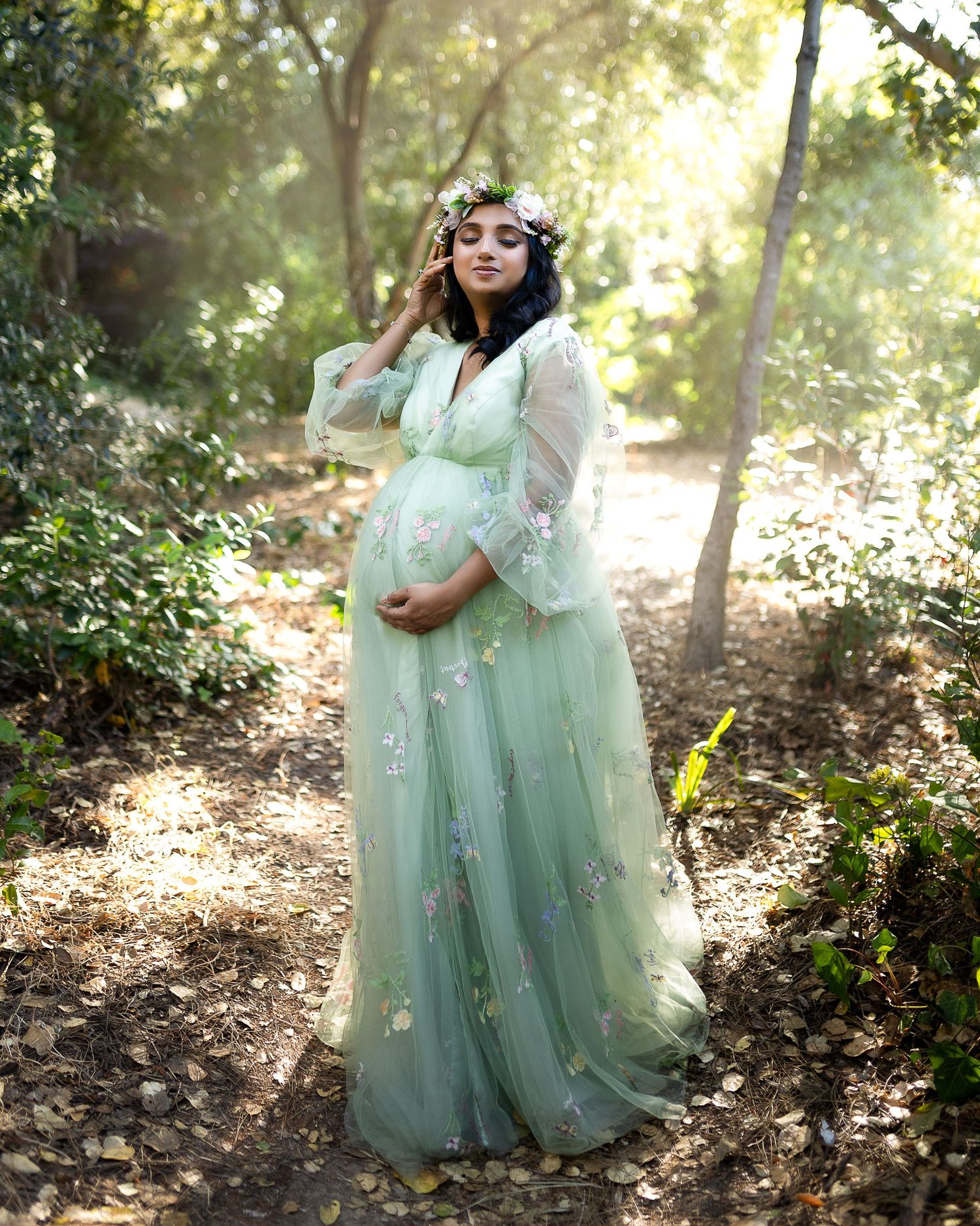 Ethereal maternity photo in a mint green tulle gown with floral crown amidst sunlit forest setting.