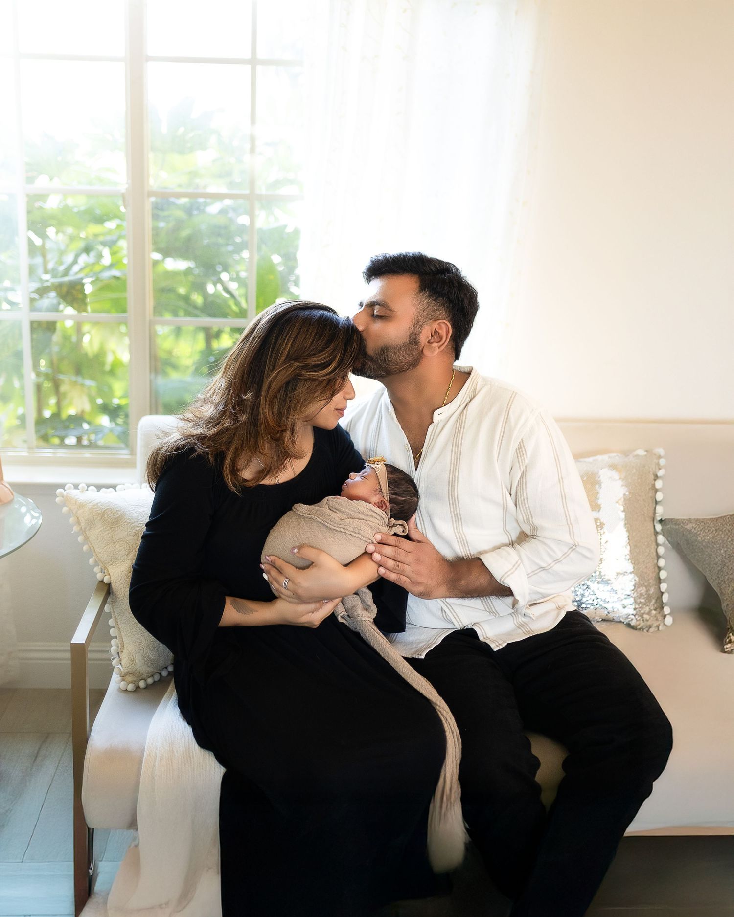 Couple sitting together on white couch near window while holding and looking lovingly at their newborn baby.