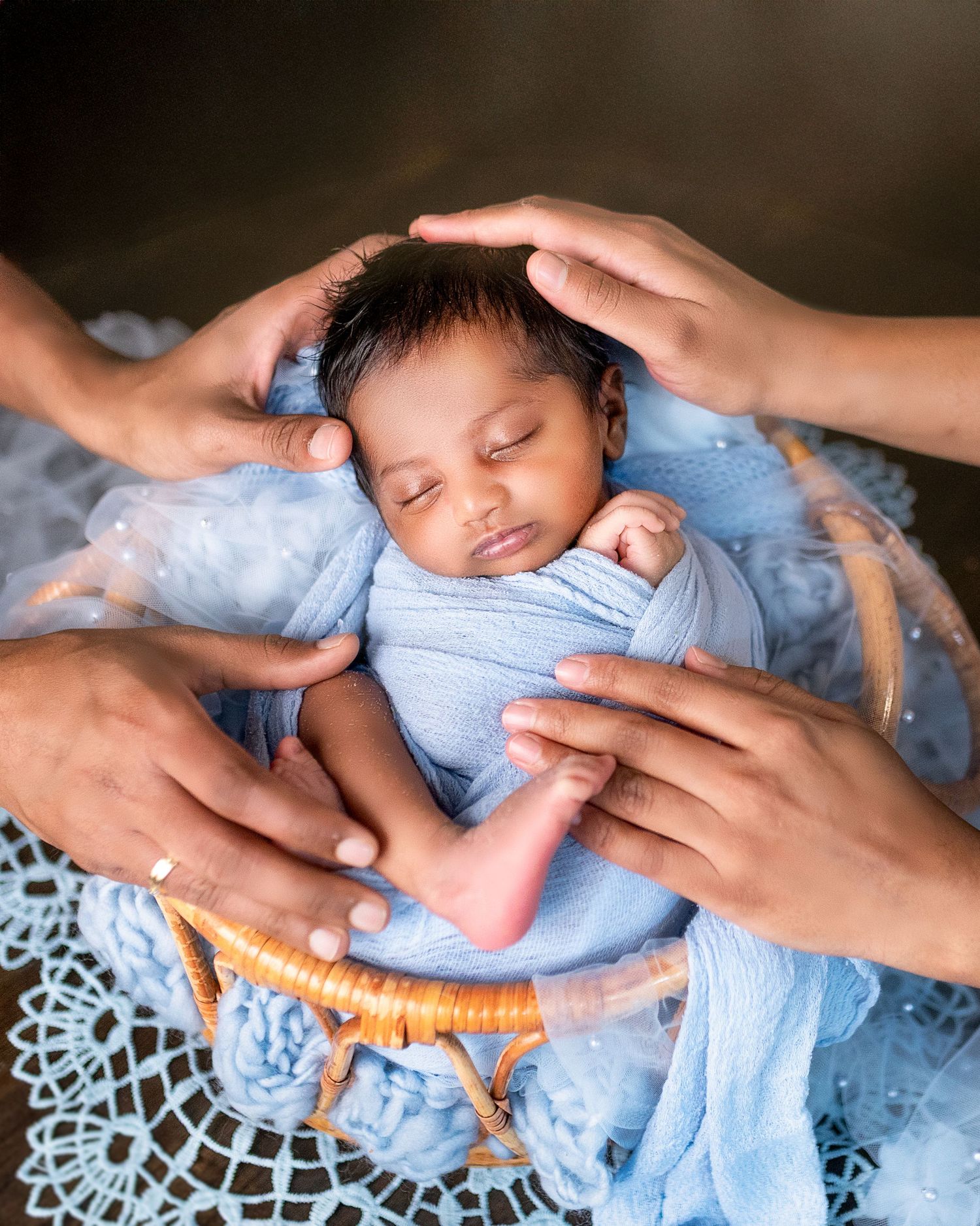 A sleeping baby wrapped in light blue fabric is gently cradled by caring hands on a delicate lace doily.