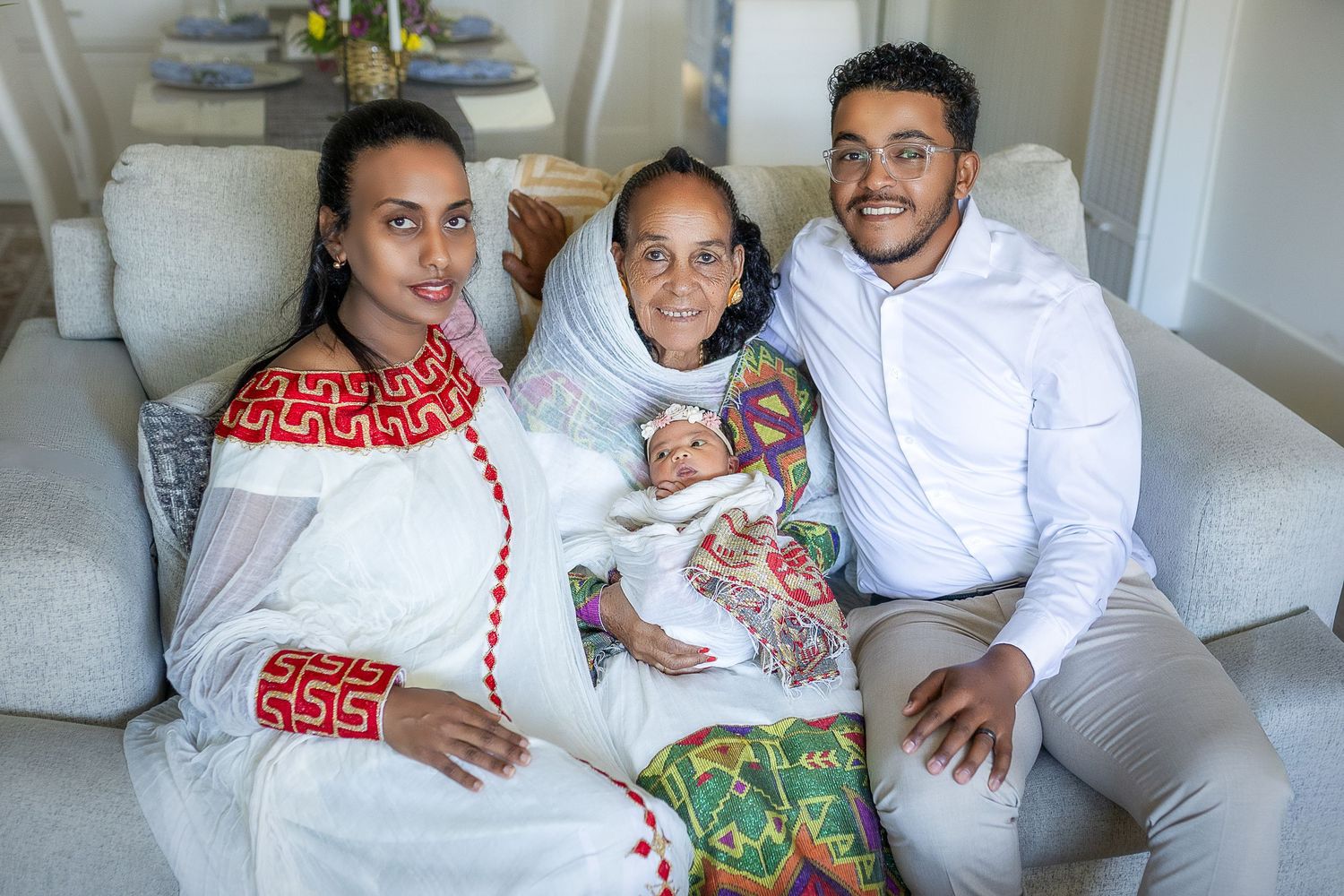 A family relaxes together on a white couch while holding a baby in a bright, modern living room setting.
