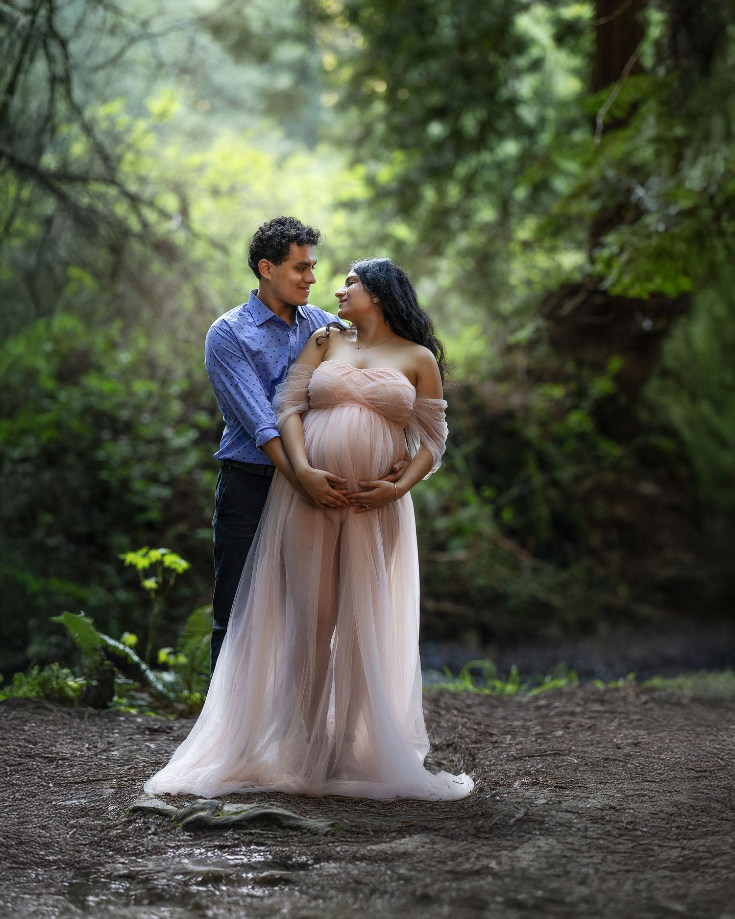 Romantic maternity portrait session showing couple embracing on a forest path surrounded by lush greenery.