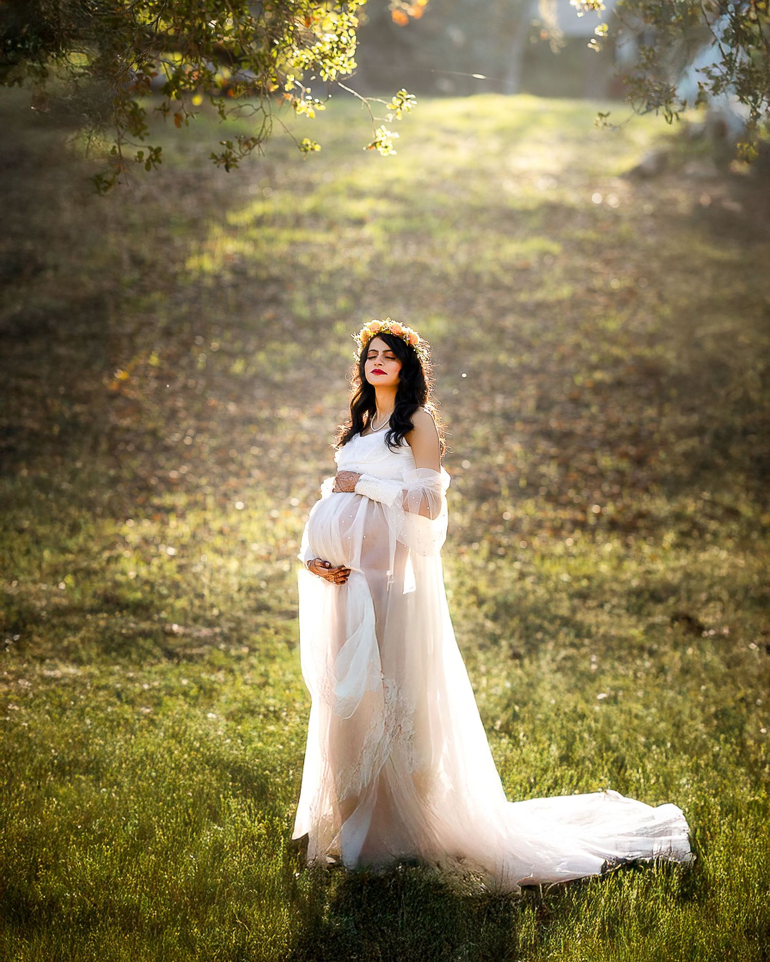 A couple in elegant white attire embraces while standing in a sunlit grassy meadow during golden hour.