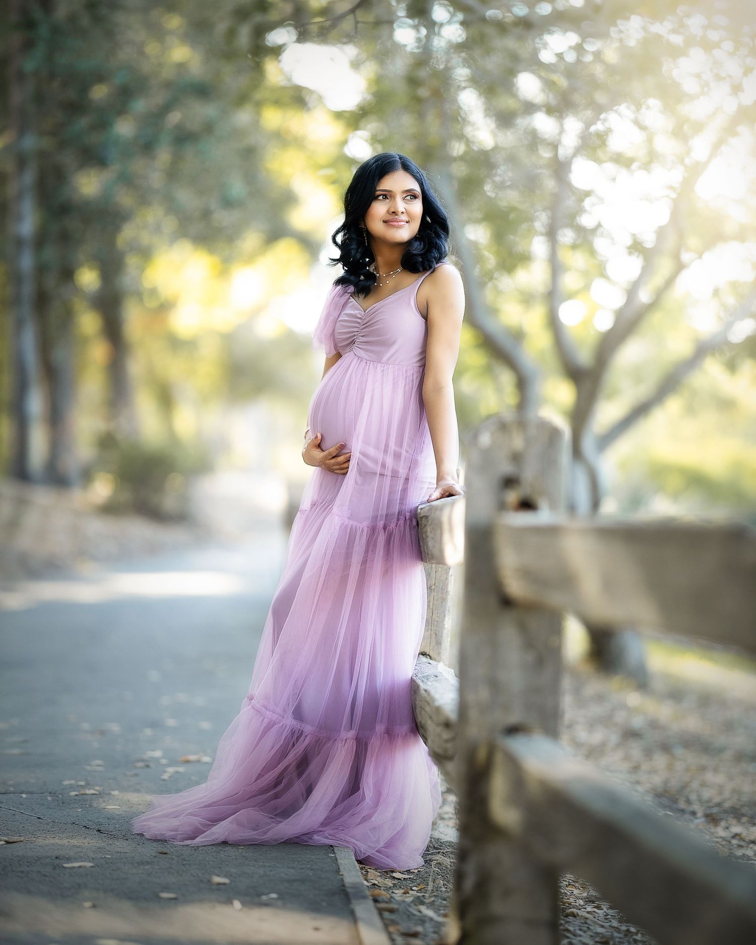 A person in a flowing lilac tulle dress poses gracefully in a sunlit outdoor setting with trees in the background.