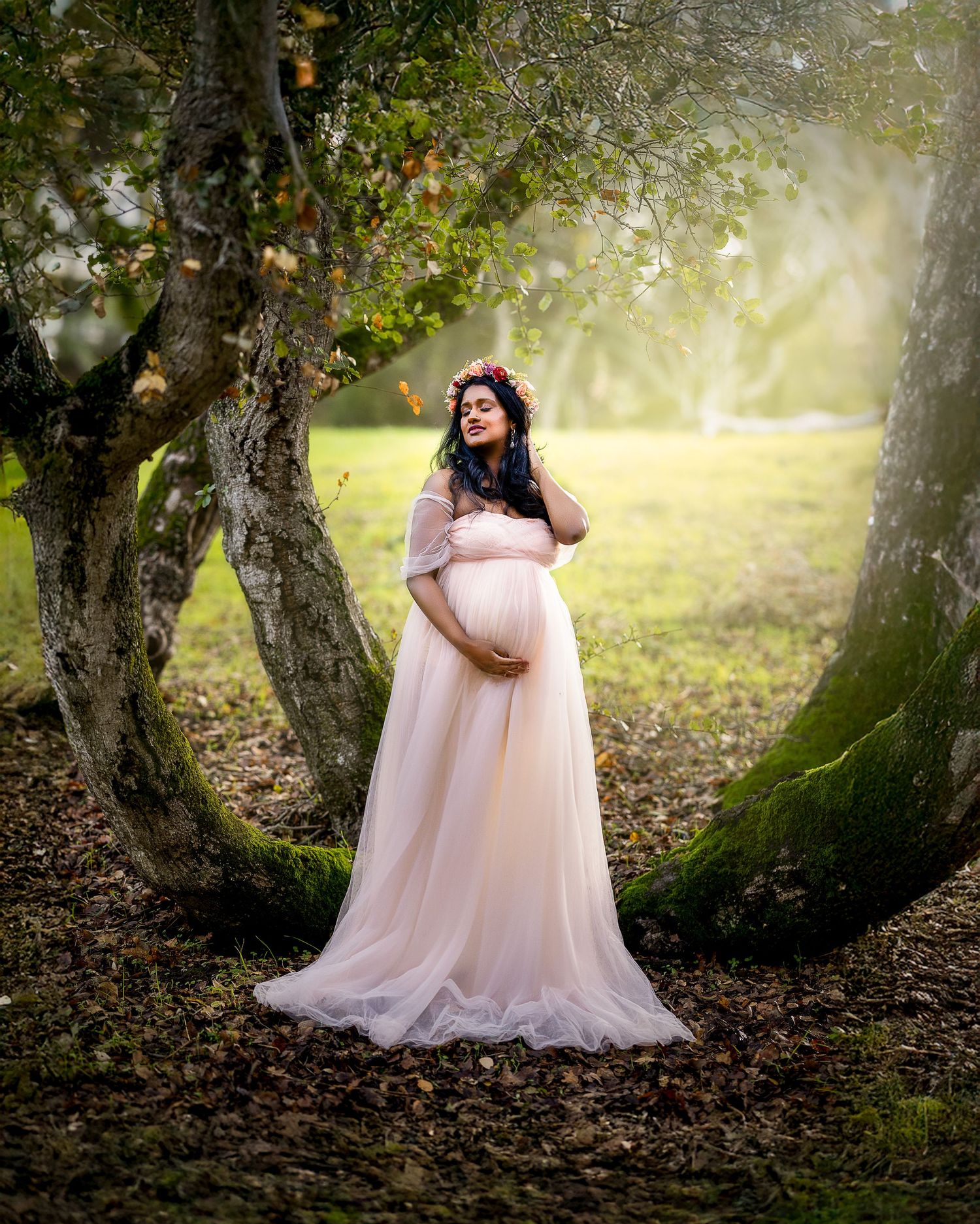 A figure in a flowing pink tulle gown poses gracefully among mossy trees in an ethereal outdoor setting.