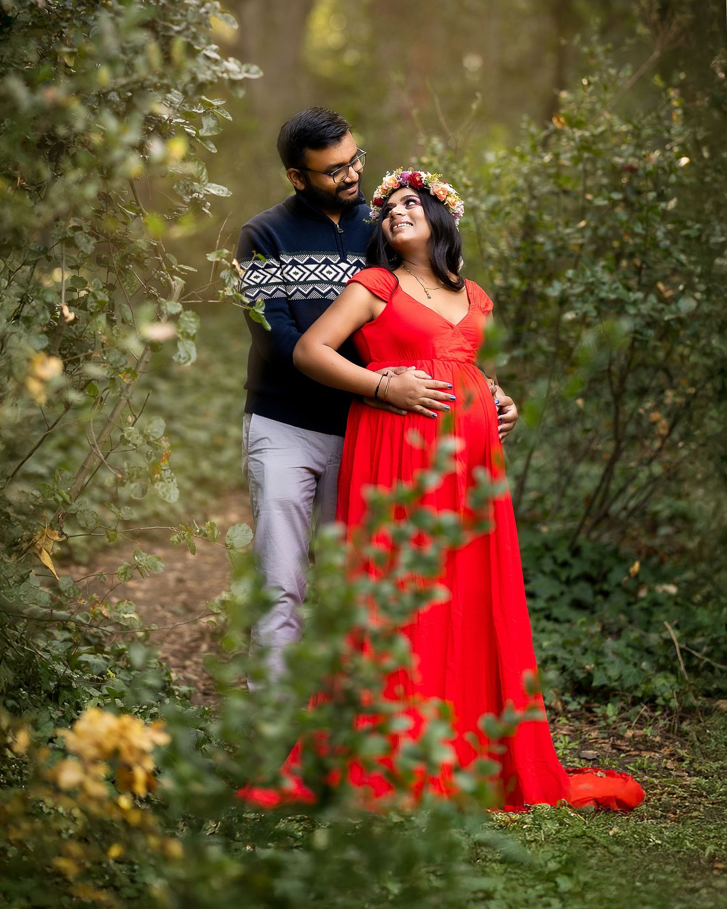 A couple in a red dress and casual attire share intimate moments in a lush garden setting with soft natural lighting.