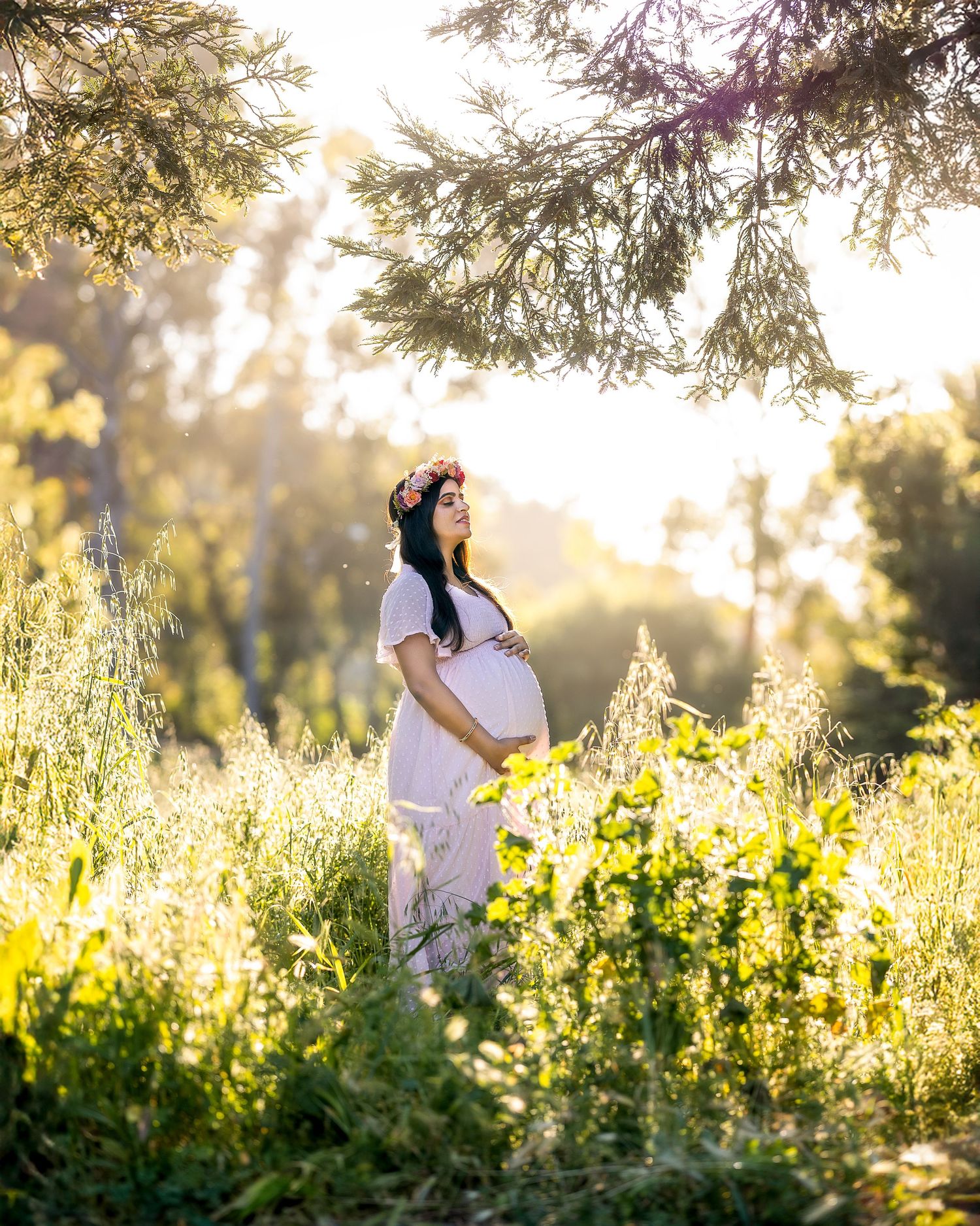 A maternity photoshoot in a sunlit meadow with yellow wildflowers and trees during golden hour.