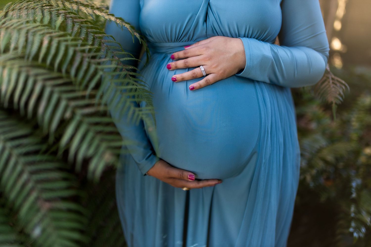 A pregnant woman in a flowing light blue dress cradles her belly while standing among lush green ferns in a garden.