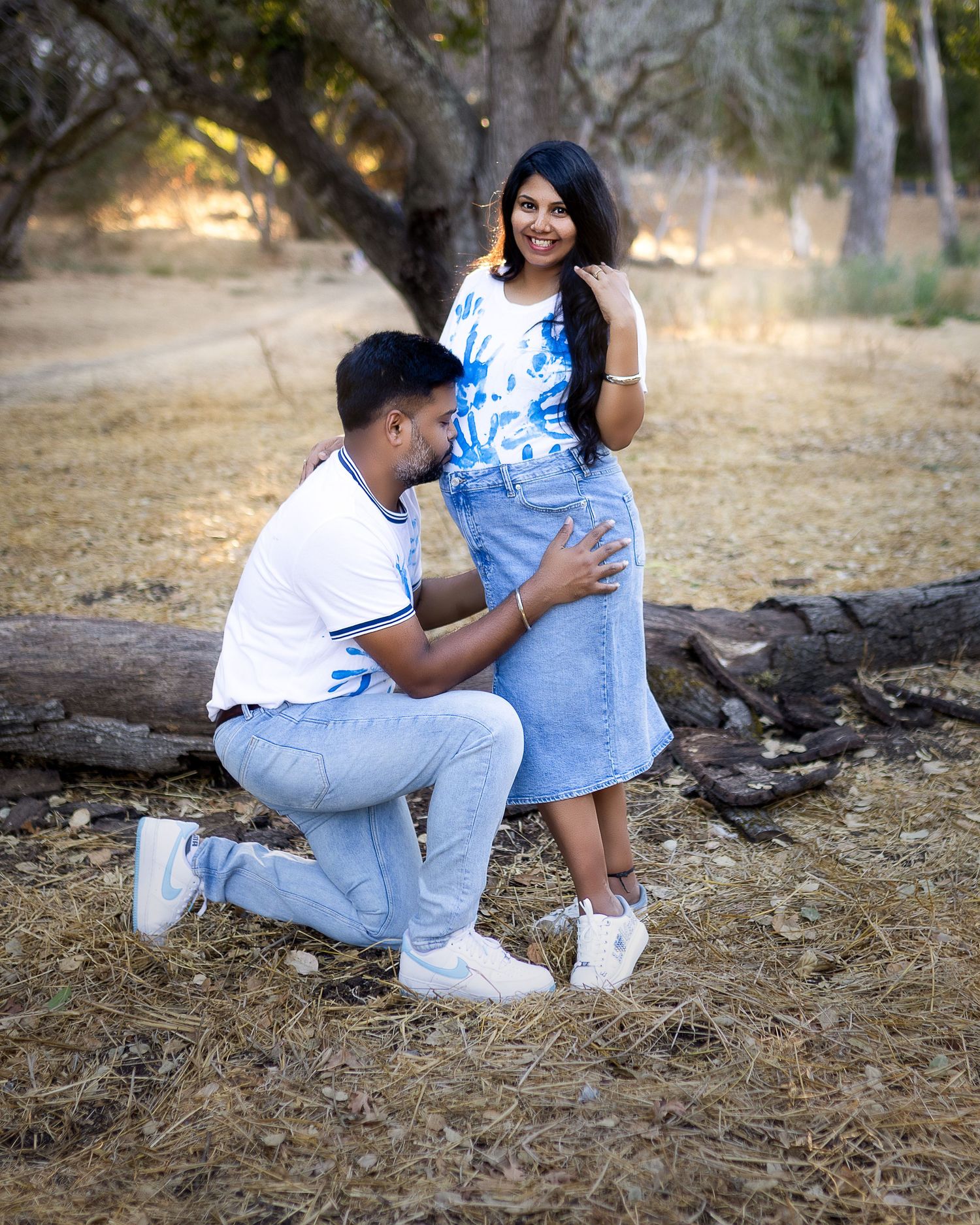 A couple in matching white and denim outfits sits together on fallen logs in a rustic outdoor setting.