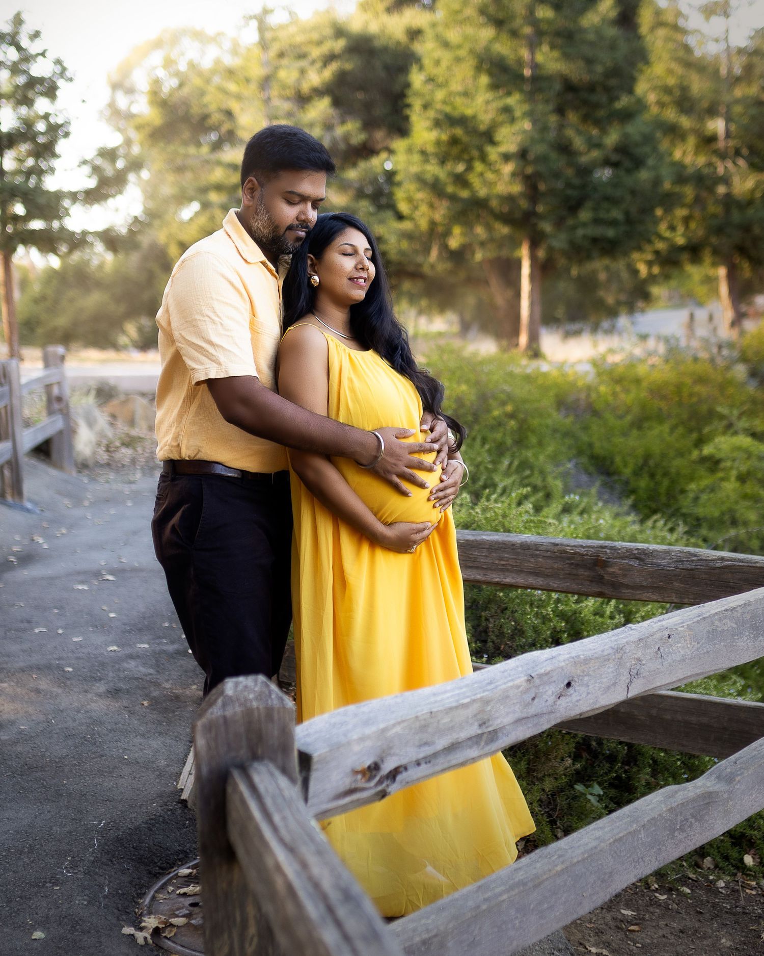 A couple embraces near a rustic wooden fence during golden hour, with one wearing a bright yellow dress.
