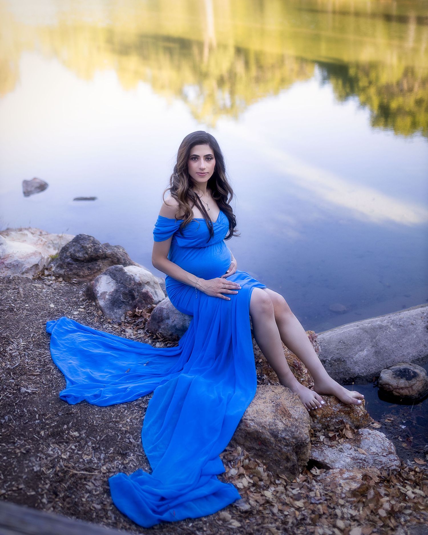 A woman in a flowing blue dress sits gracefully by a serene lake during a maternity photoshoot at sunset.