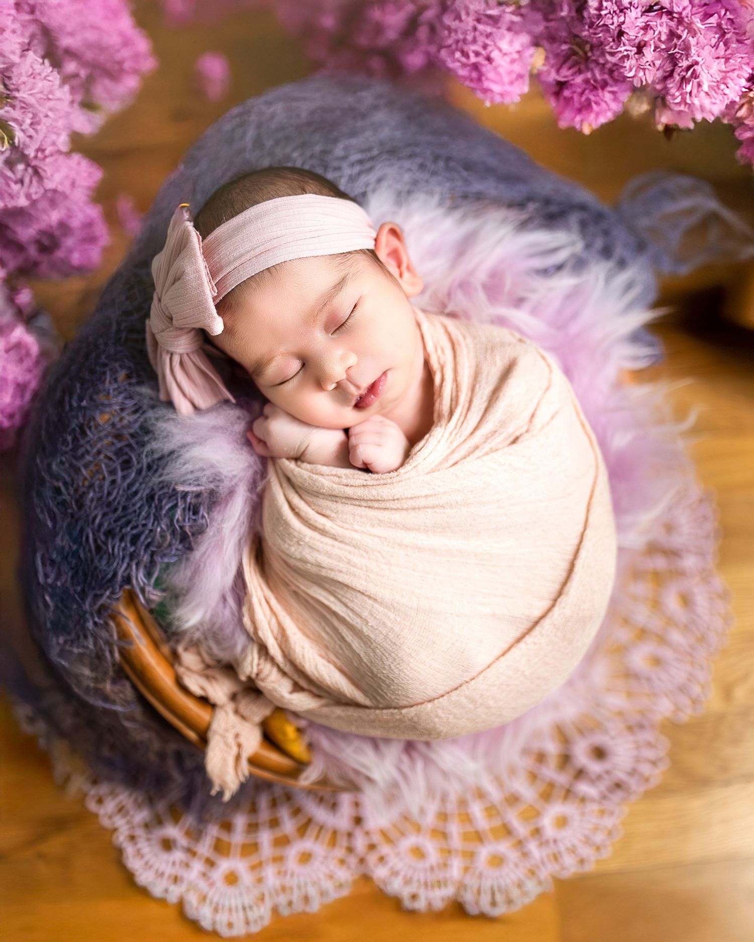 A sleeping newborn wrapped in beige fabric lying on a delicate purple lace doily against a wooden surface.