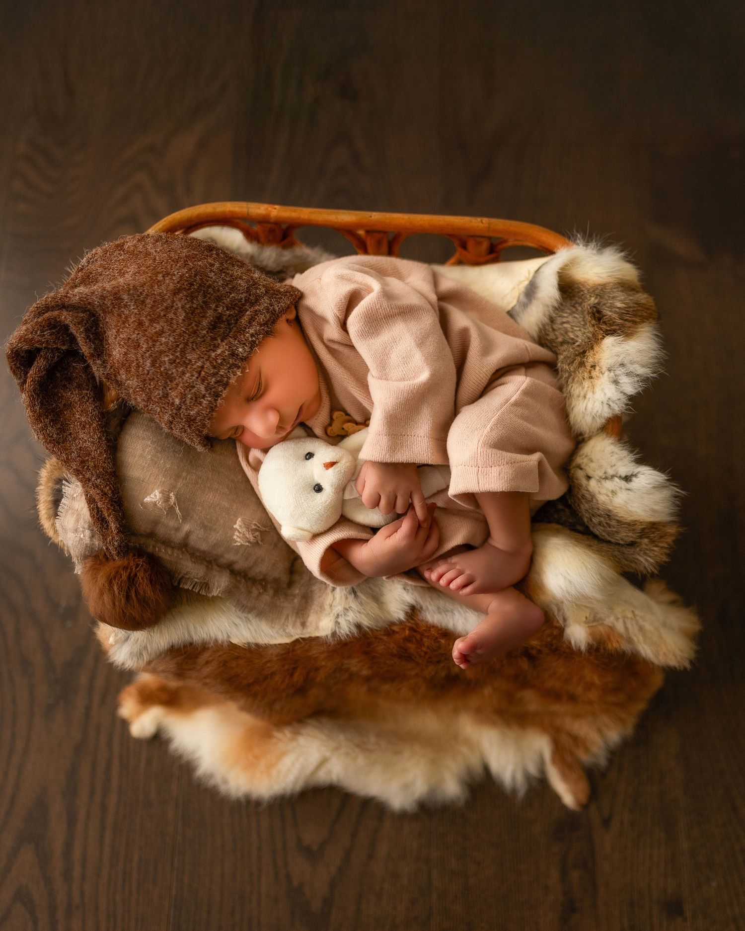 A sleeping baby wearing a pink onesie rests on soft brown and white fur blankets in a cozy studio setting.