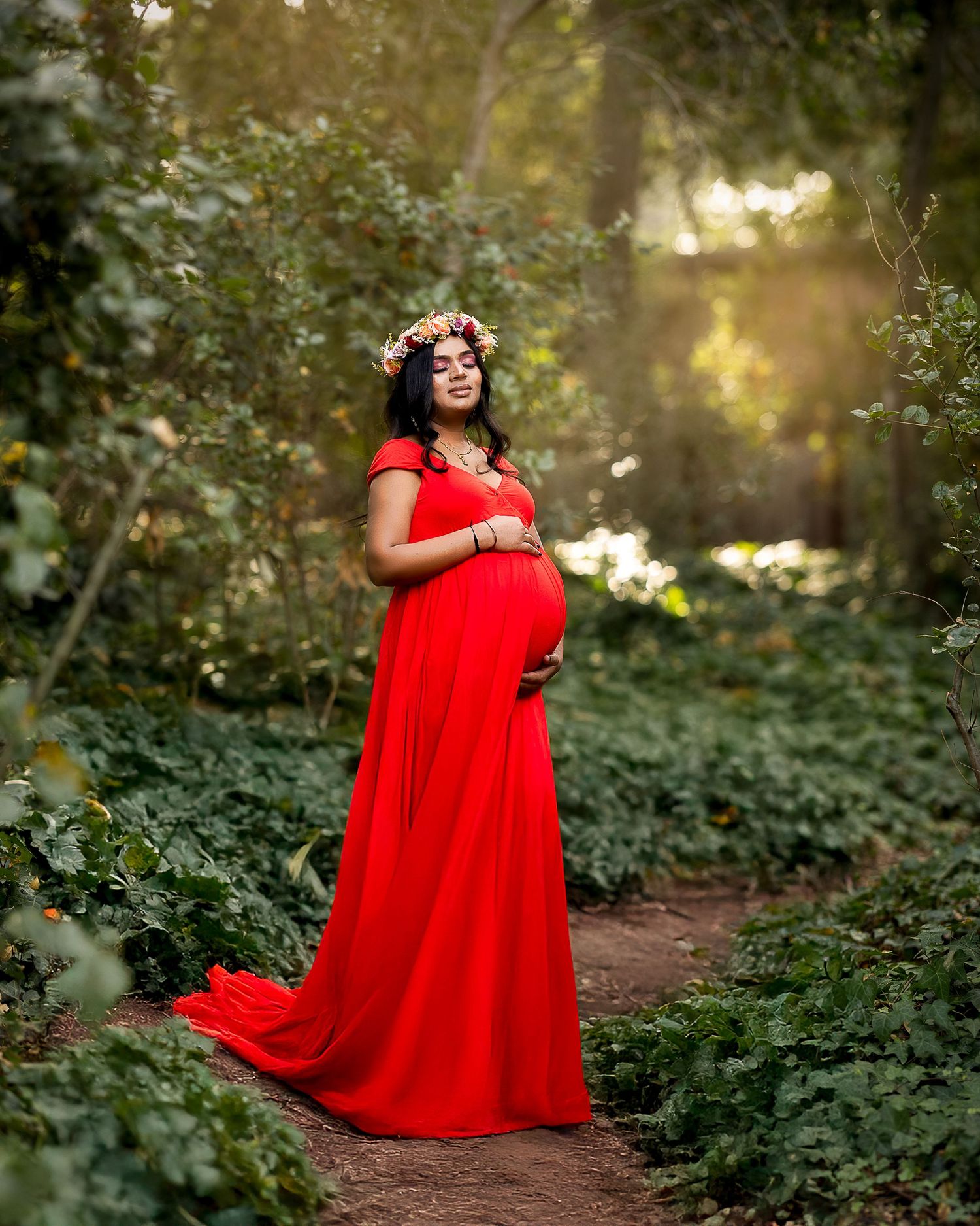 A woman in a flowing red dress poses gracefully in a sunlit forest setting during a photo session.
