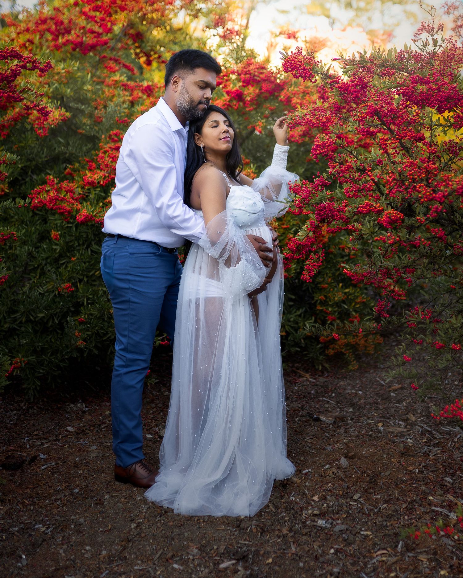 A romantic couple poses in front of vibrant red flowering bushes during a photo session.