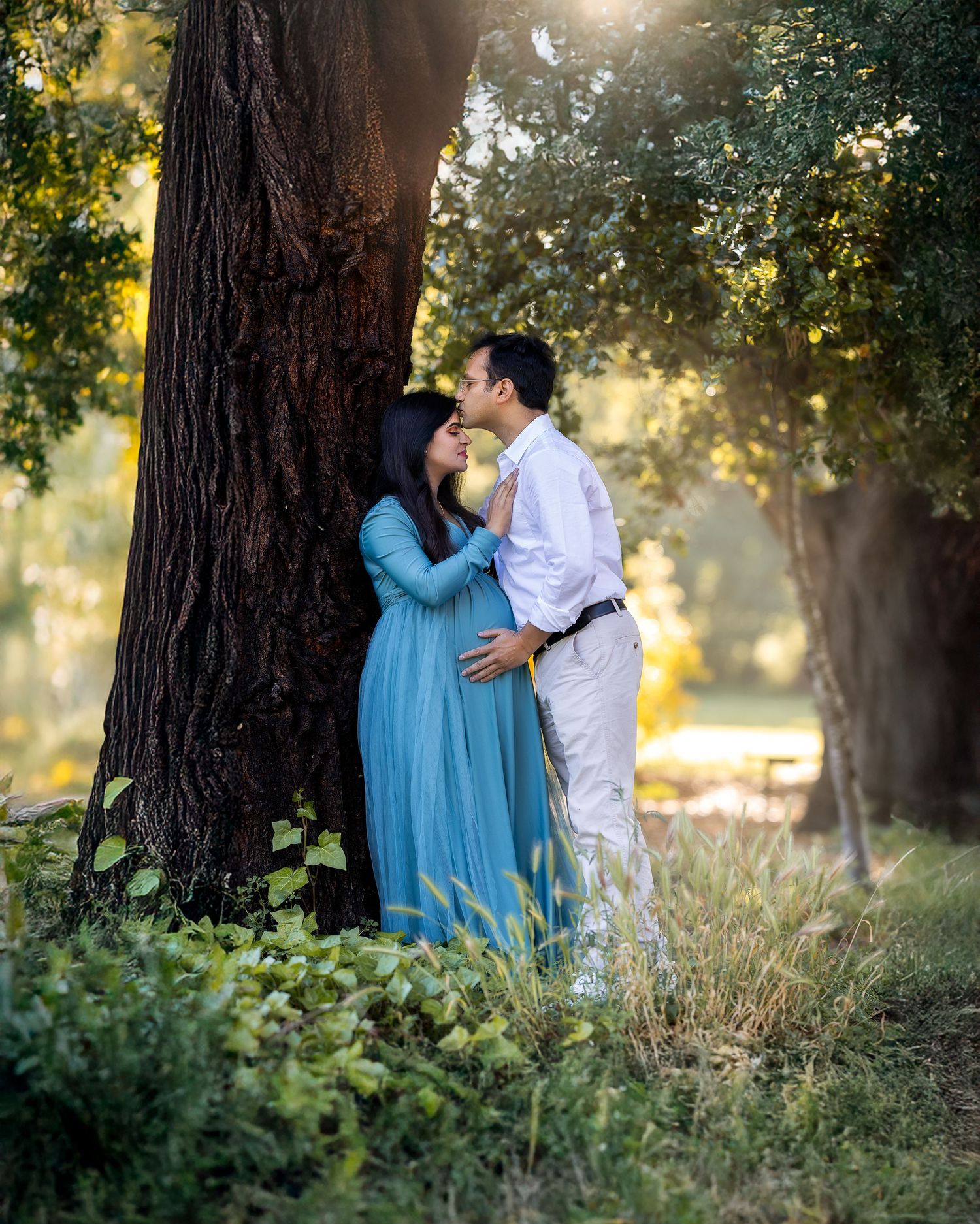 Two people share a tender moment in a sunlit forest setting with tall trees and golden light filtering through.