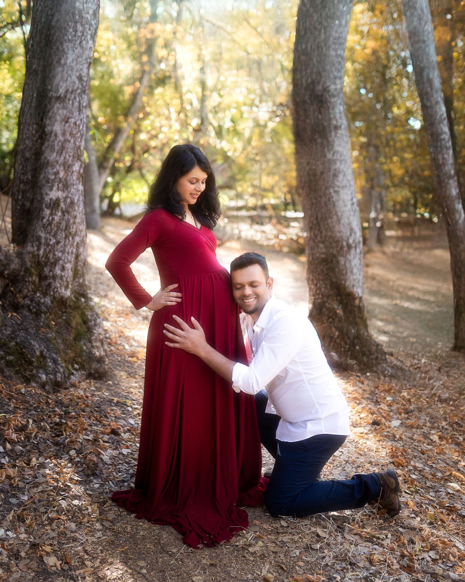 A couple poses for a maternity photo in a forest setting, with the woman wearing an elegant red gown.
