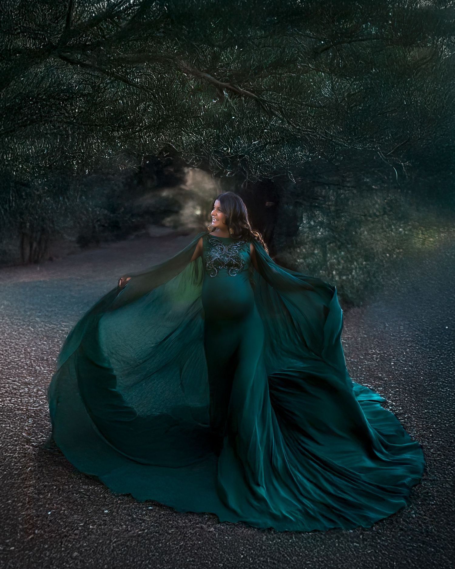A dark emerald gown billows dramatically across a shadowy forest floor at dusk.