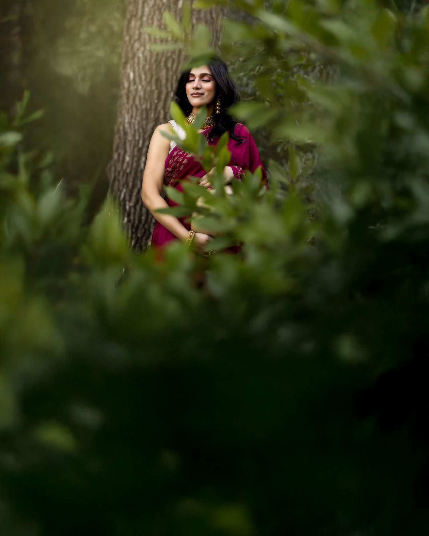 A figure in a burgundy dress poses gracefully among lush green foliage in an artistic outdoor portrait session.