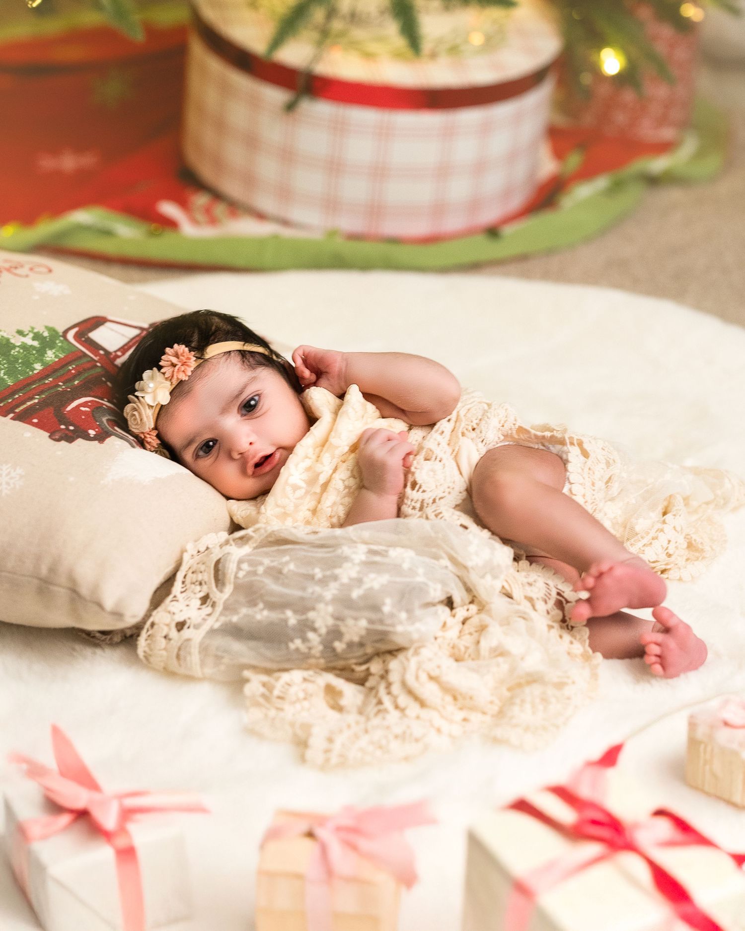 Sleeping baby in delicate white lace dress with pink ribbons and Christmas decorations in background.