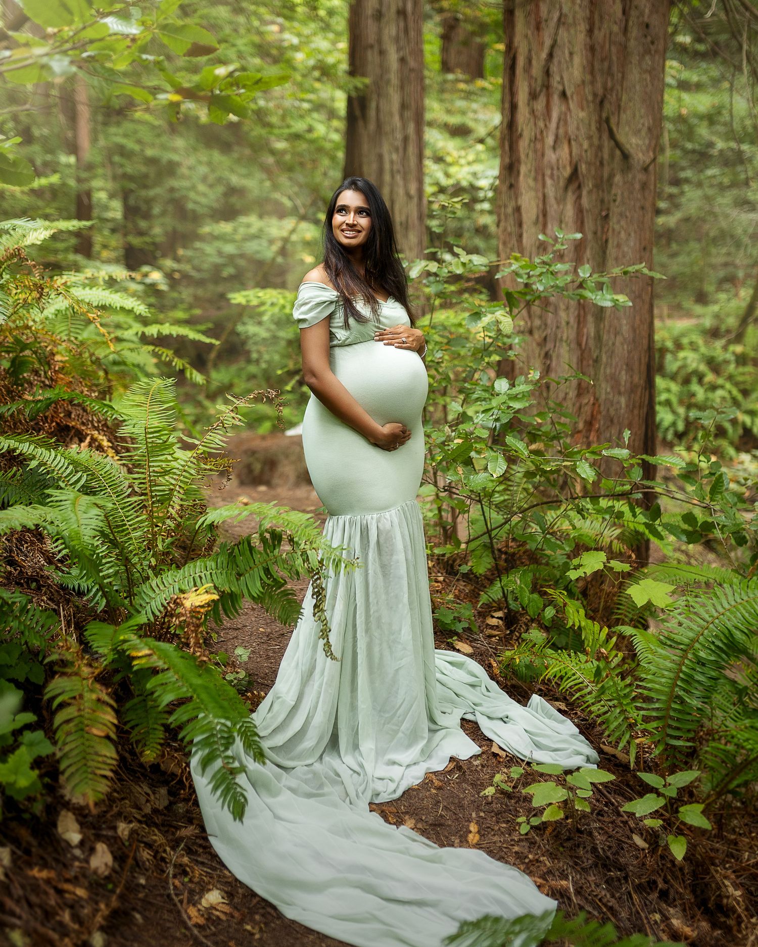 A person in a mint green flowy dress poses gracefully among lush ferns in a serene redwood forest setting.