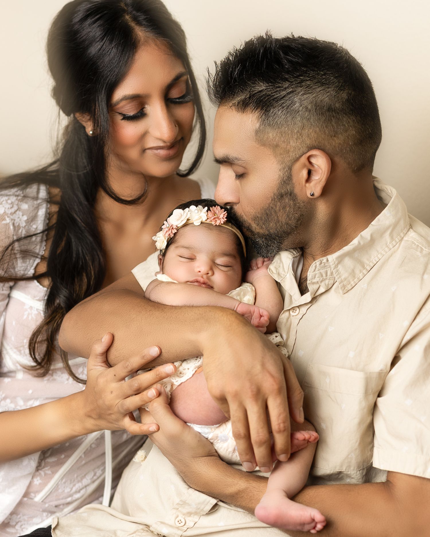 A tender family moment as parents cradle their sleeping baby wearing a floral headband in a warm studio portrait.