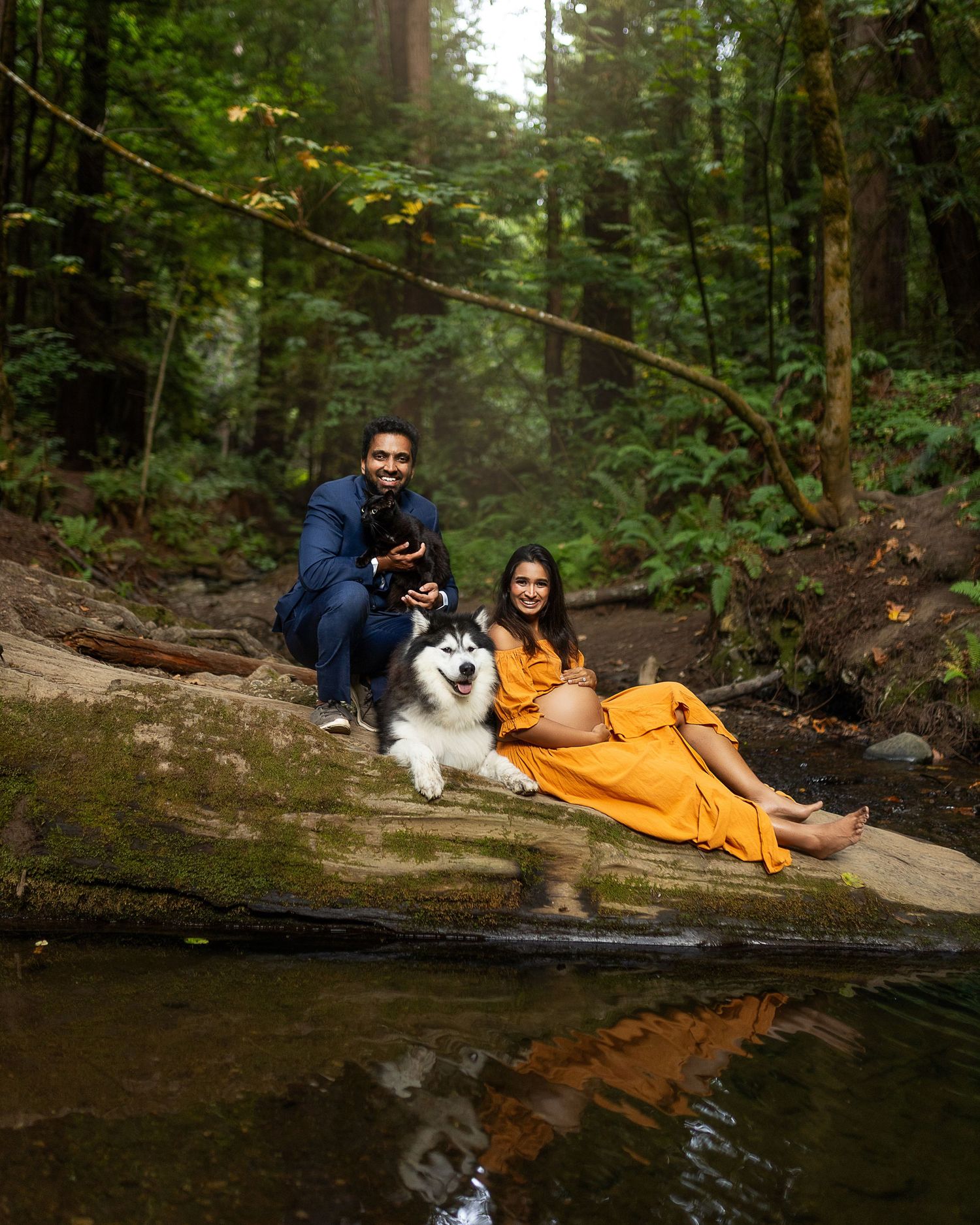 A couple and their fluffy white dog relax on a large rock by a serene forest stream on an autumn day.