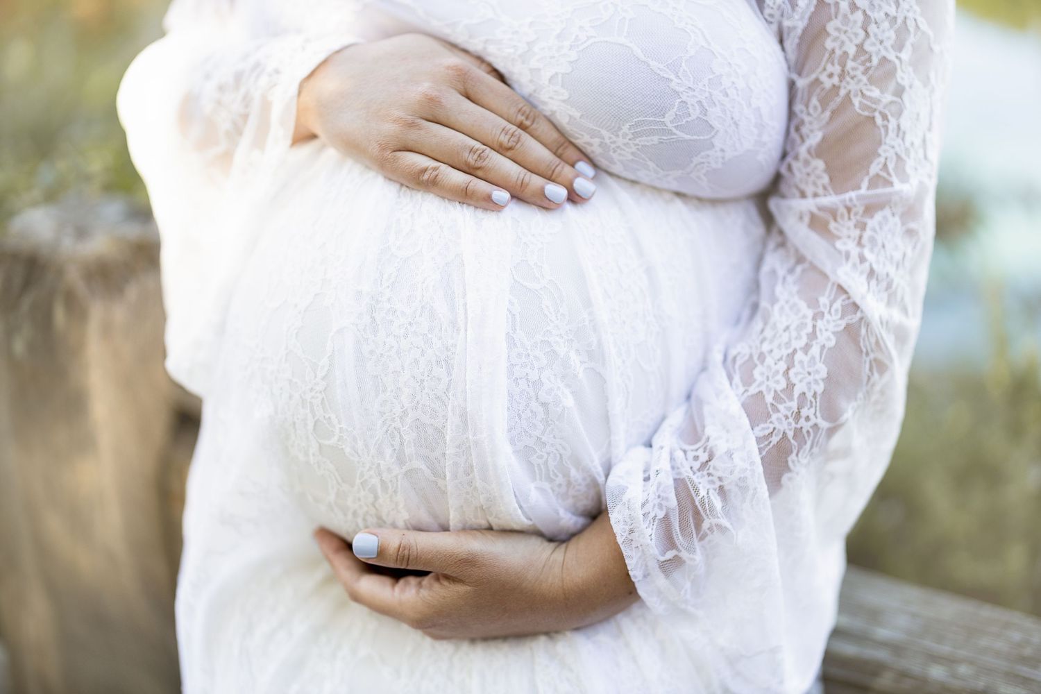 A person in a white lace dress cradles their pregnant belly while standing outdoors in natural light.