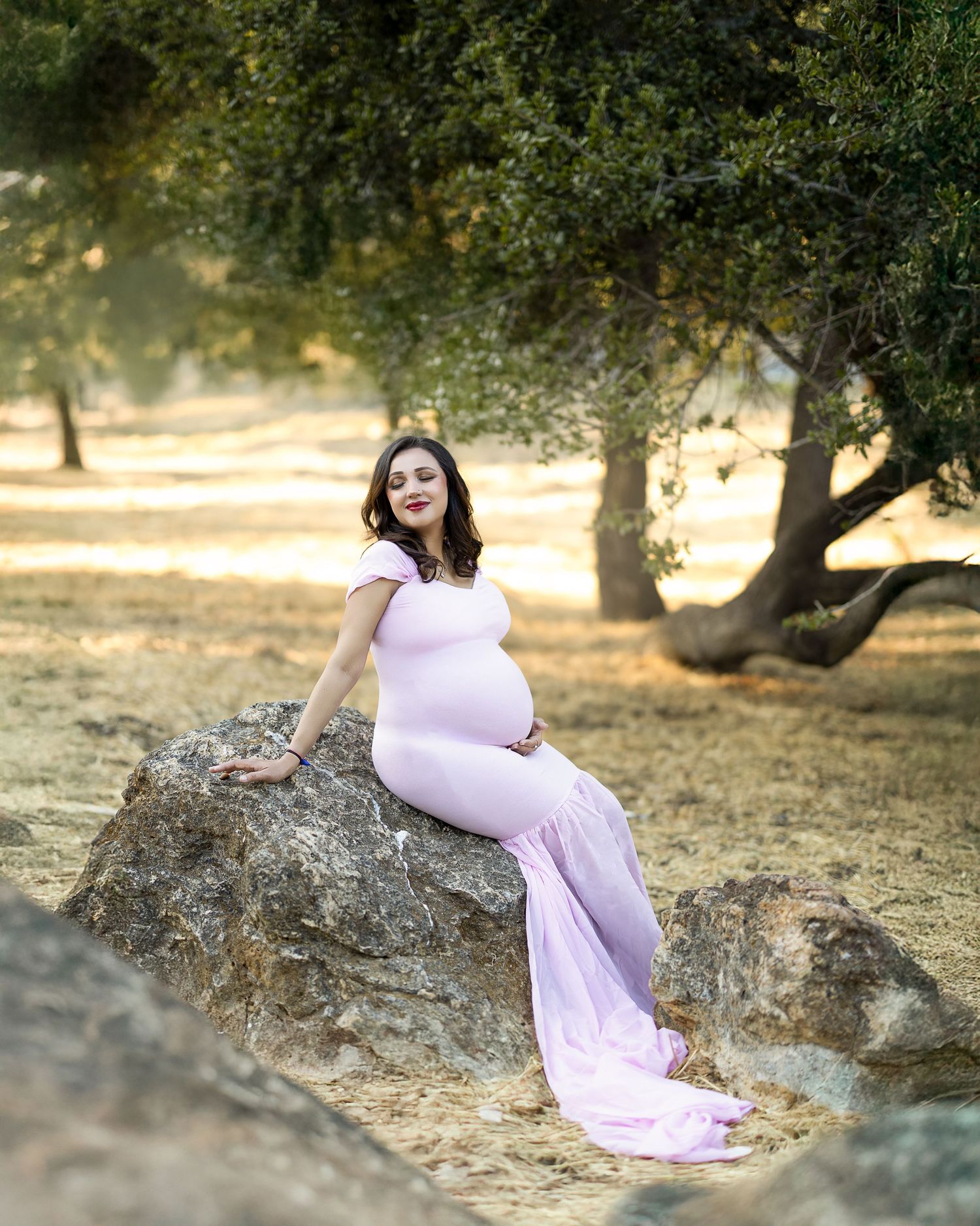 A person in a flowing lavender dress sits gracefully on a large boulder among olive trees at sunset.