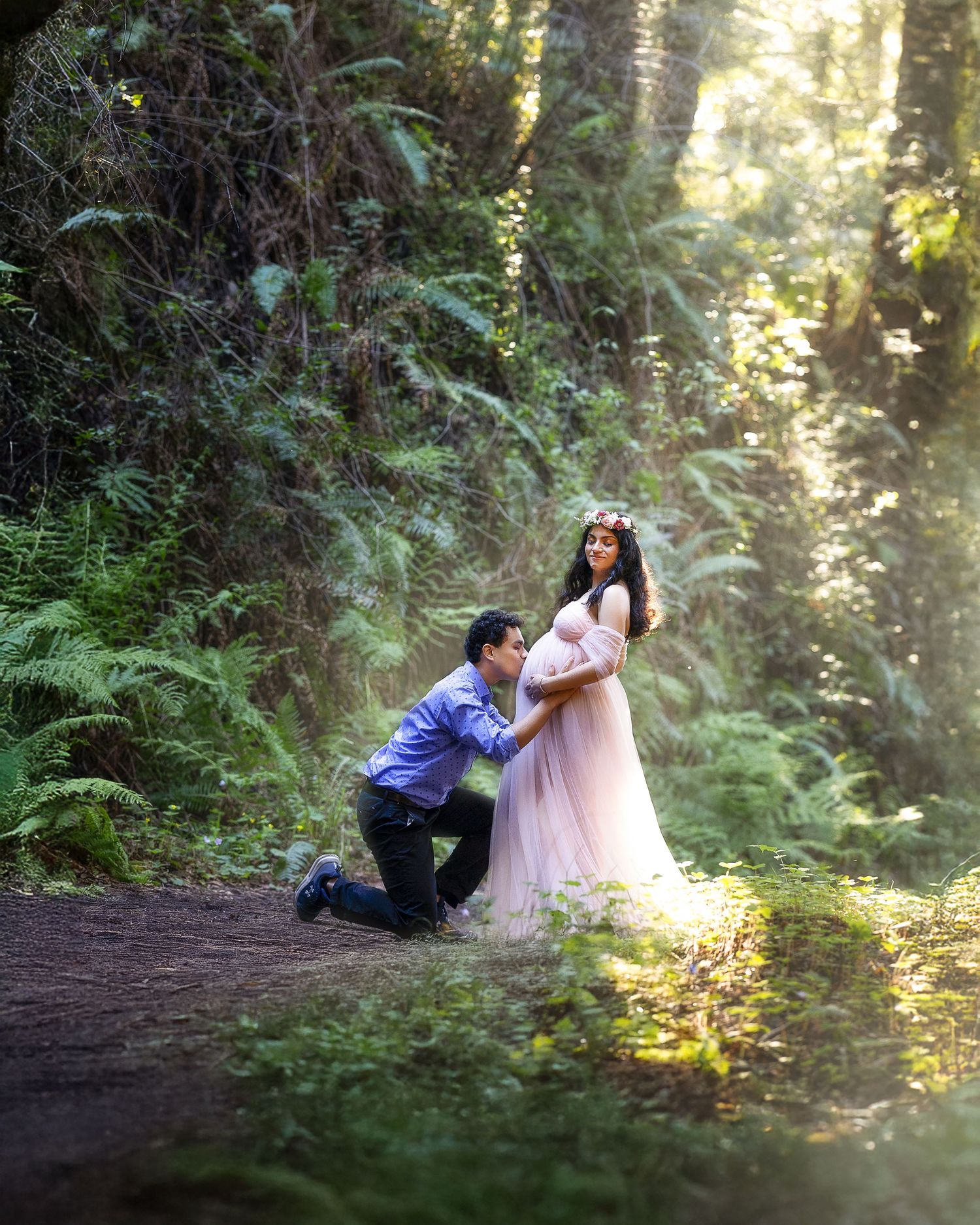 A romantic scene in a sunlit forest with a couple in formal attire sharing a tender moment on a dirt path.