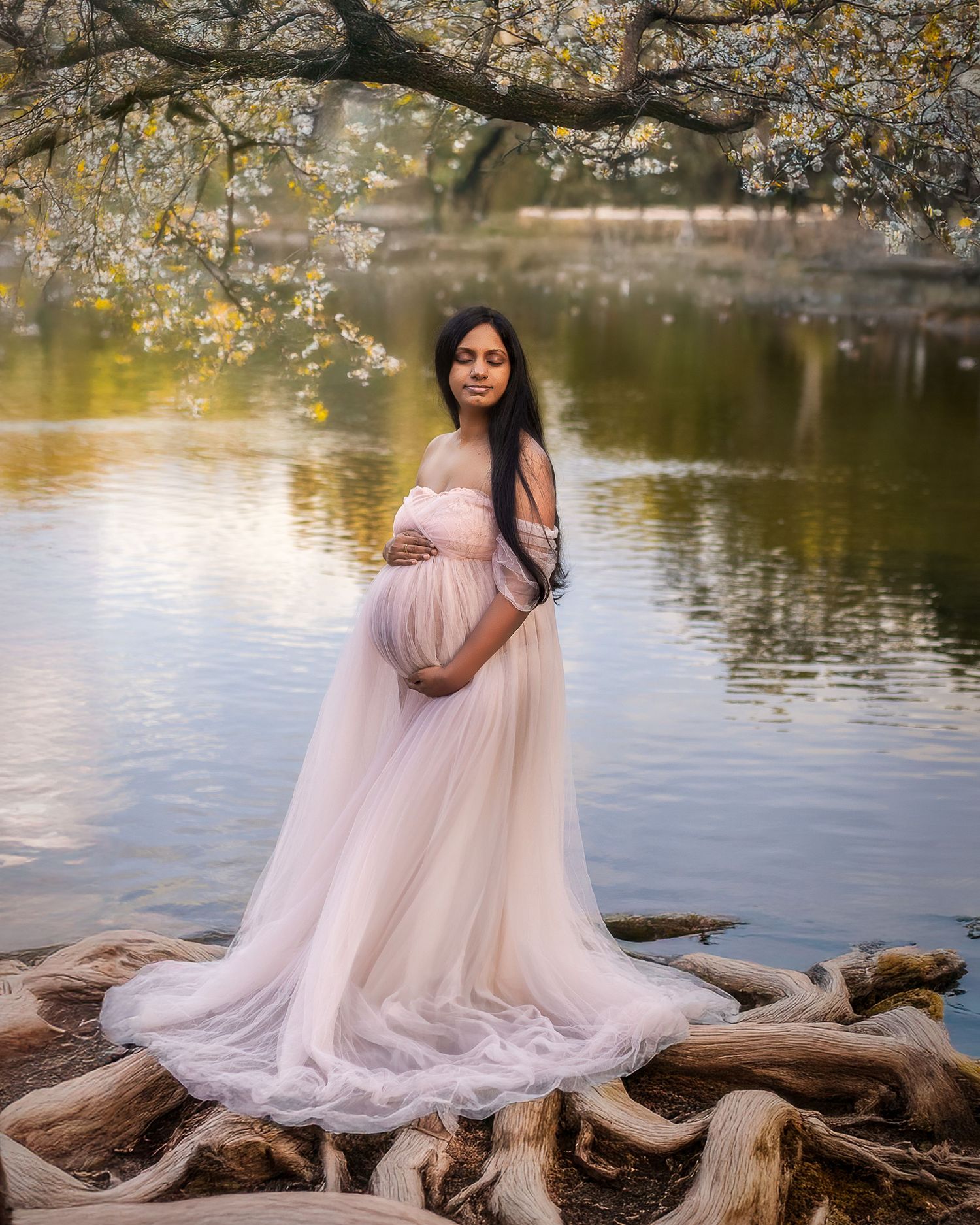 A flowing pink tulle maternity gown creates an ethereal scene by a tranquil pond with blooming spring branches overhead.