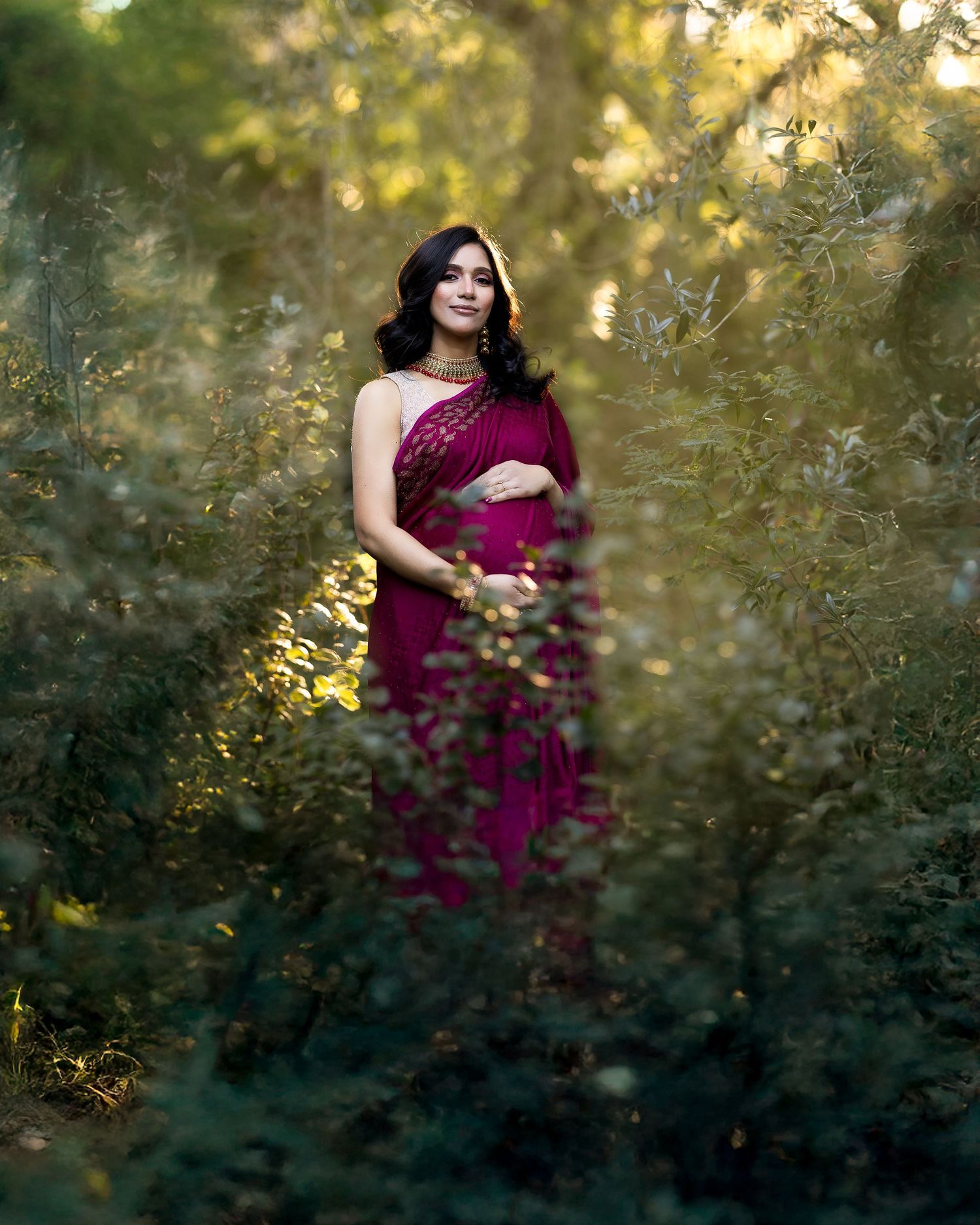 Ethereal portrait in a burgundy gown surrounded by green leaves and dappled sunlight.