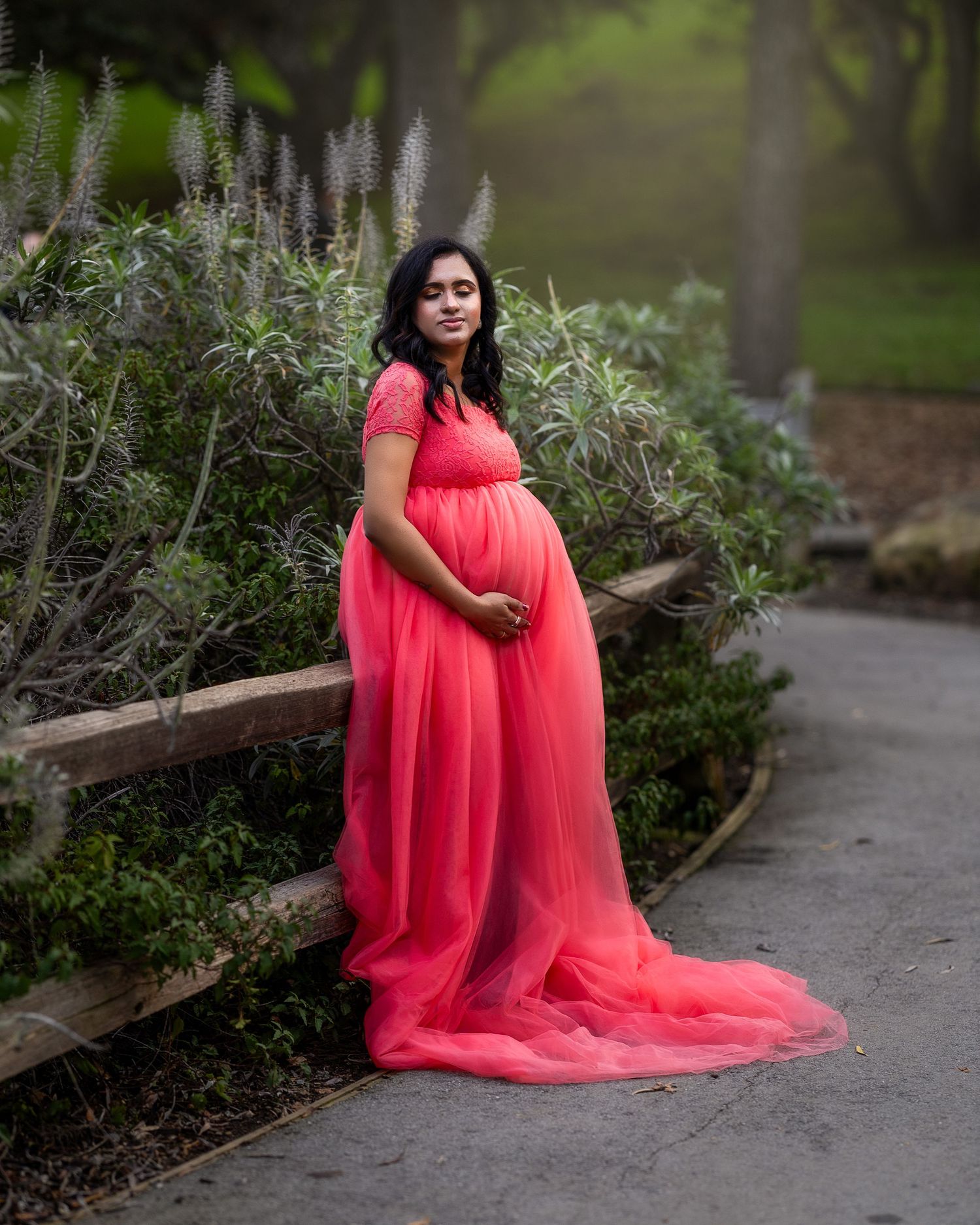 A pregnant woman in a flowing coral gown stands on a garden path surrounded by lush greenery and flowering plants.
