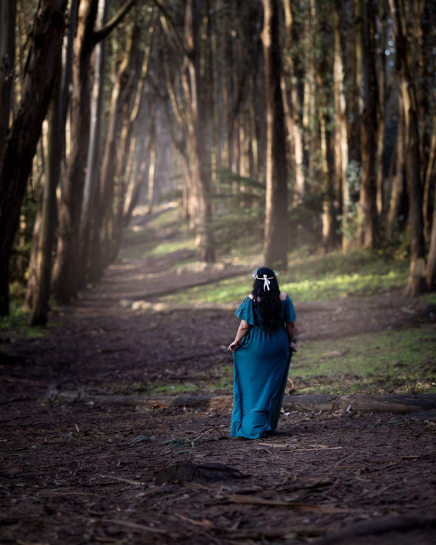 A figure in a flowing blue dress walks along a mystical forest path lined with tall trees.