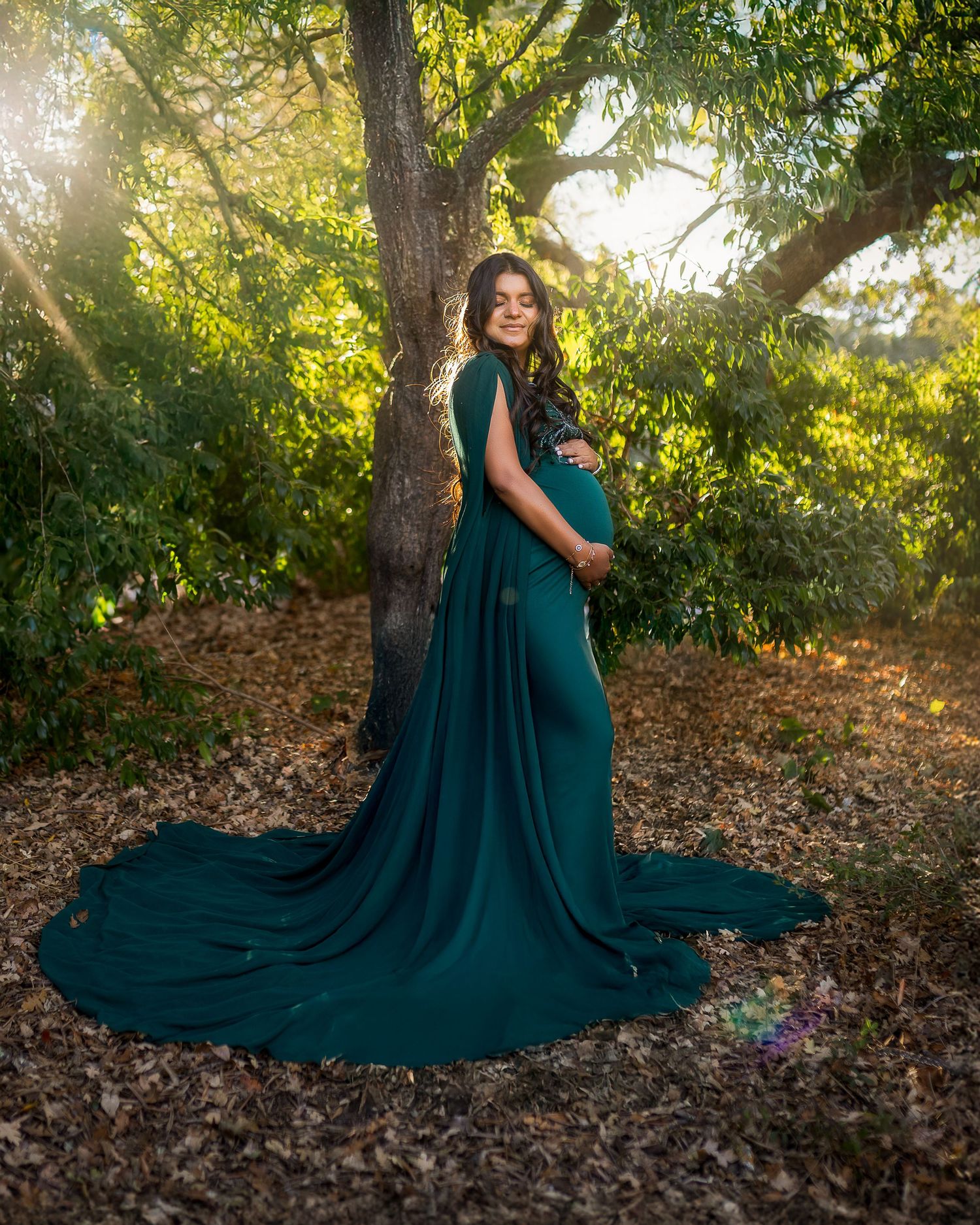 A flowing emerald green dress cascades over autumn leaves in a sunlit forest setting during a dreamy photoshoot.