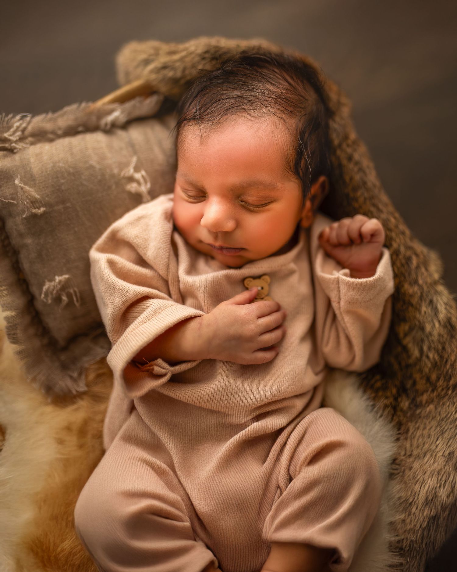 A sleeping newborn baby wearing a beige outfit lies on soft brown fur in a professional studio portrait.