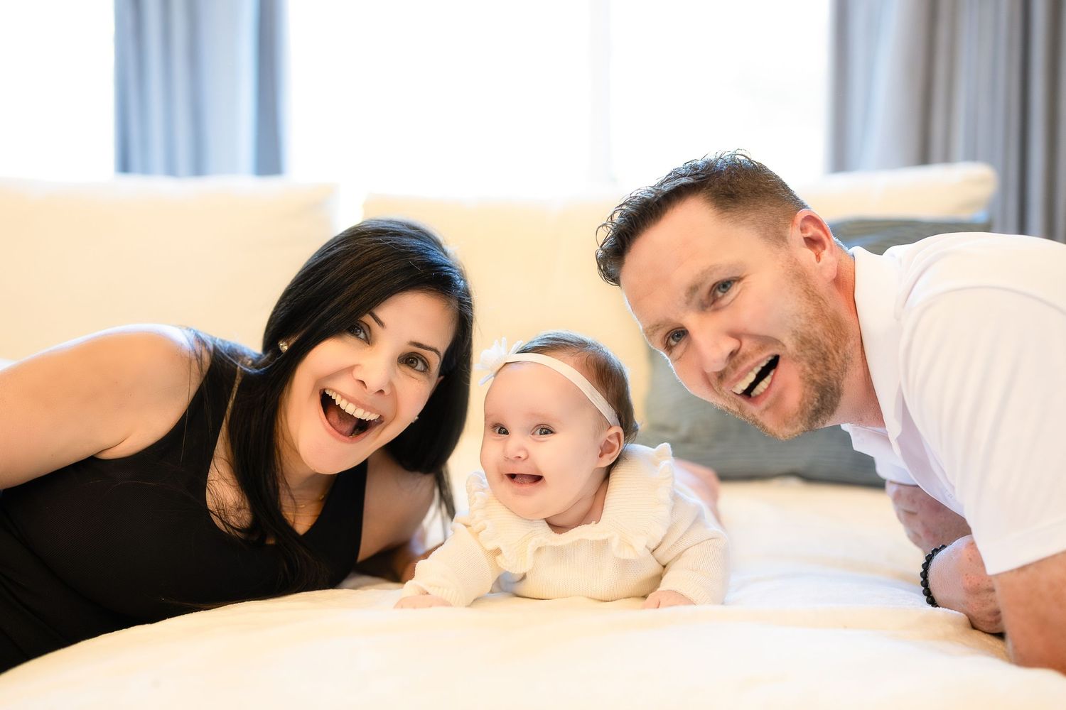A happy family lies together on a white bed, making silly faces and sharing a joyful moment.