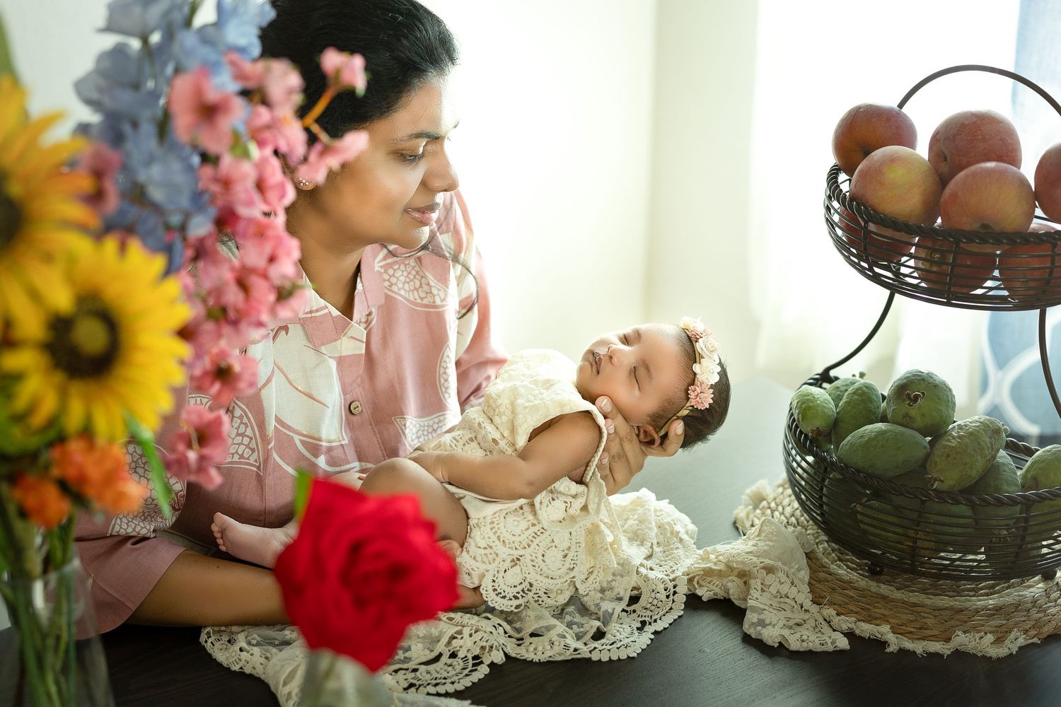 A vintage-styled scene with a baby in lace dress on table near sunflowers and fruit bowls in soft natural light.