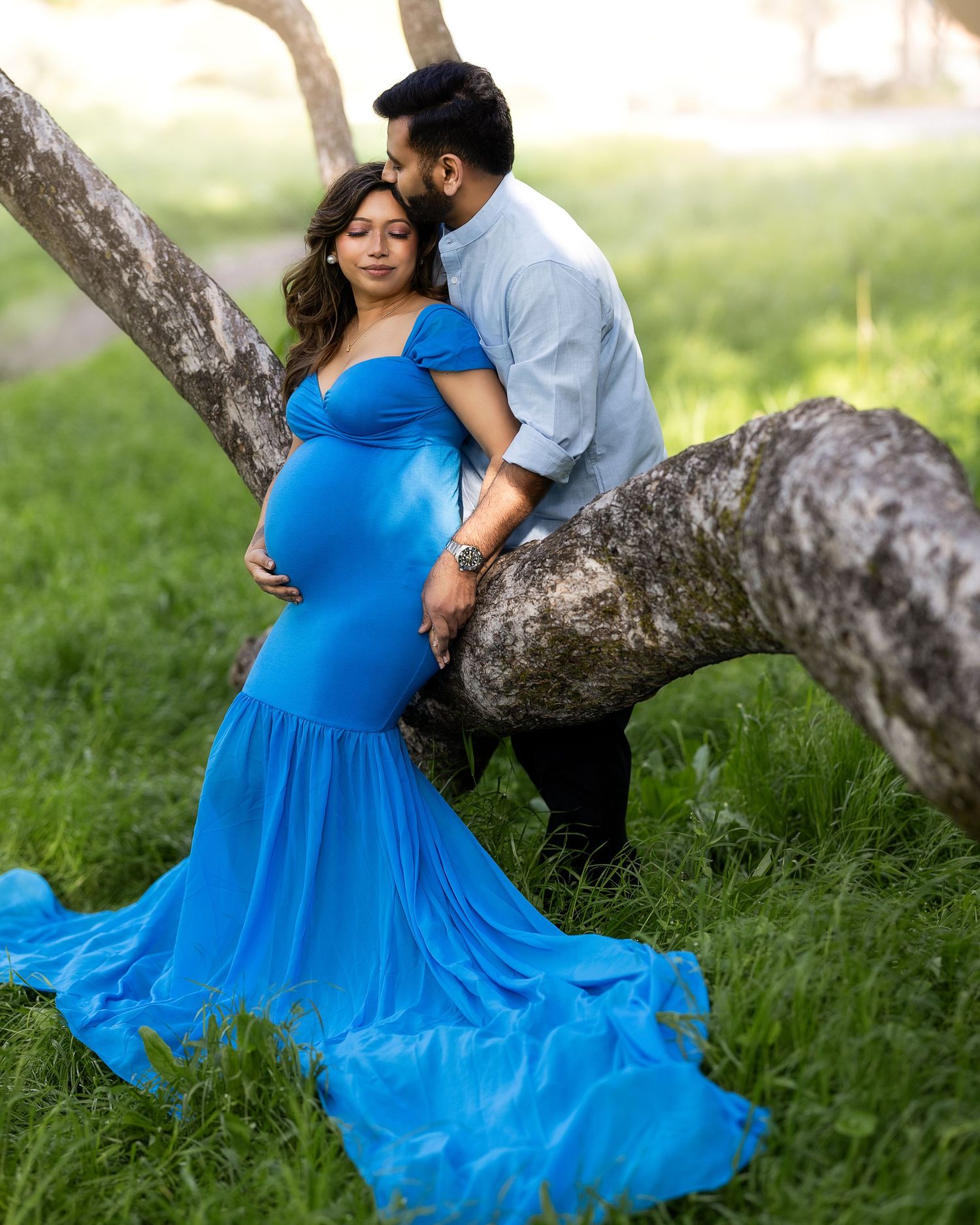 A couple shares a romantic moment under a tree in a park, with the woman wearing a flowing bright blue dress.