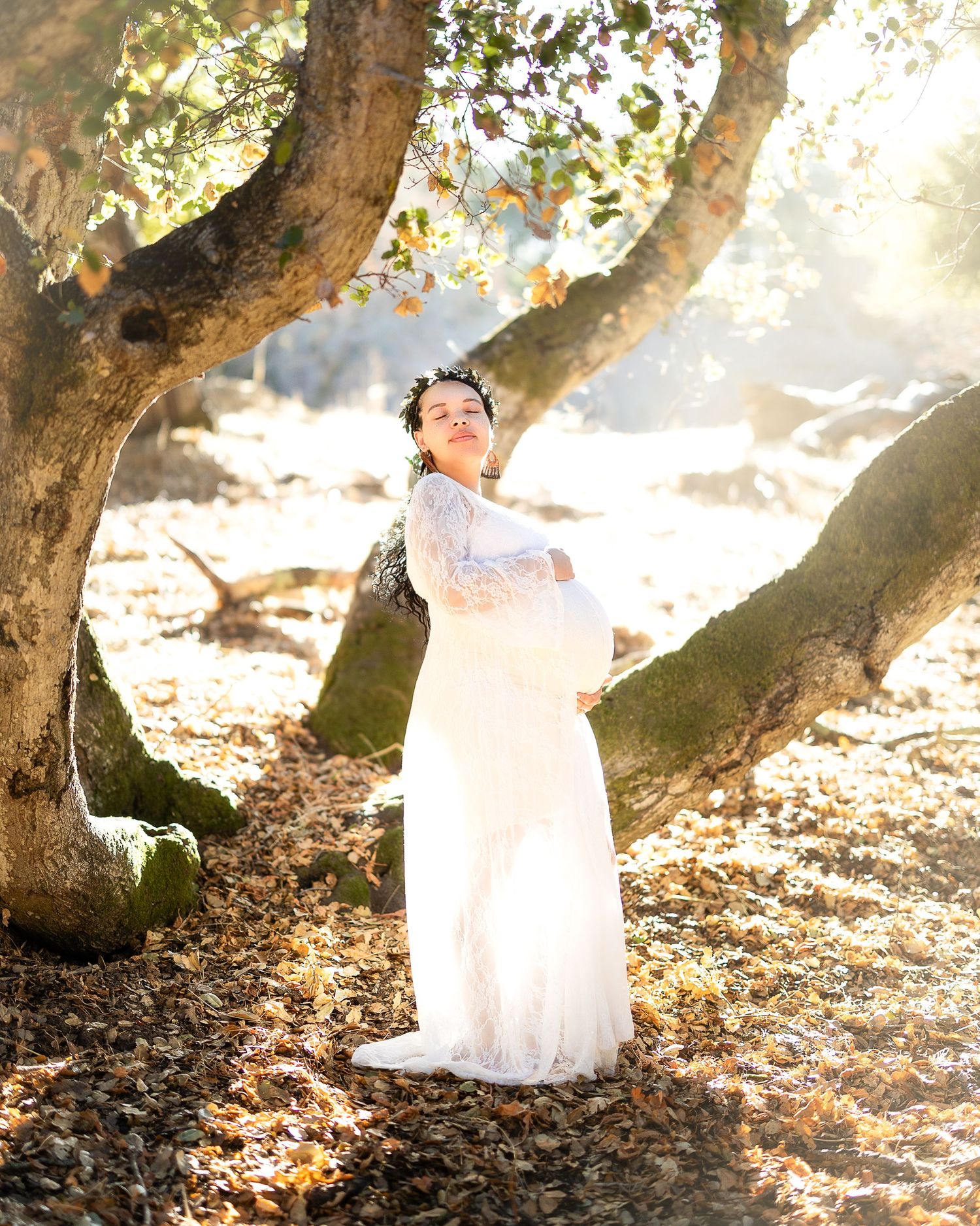 A white lace maternity dress glows in ethereal sunlight beneath autumn trees in a dreamy outdoor photoshoot.