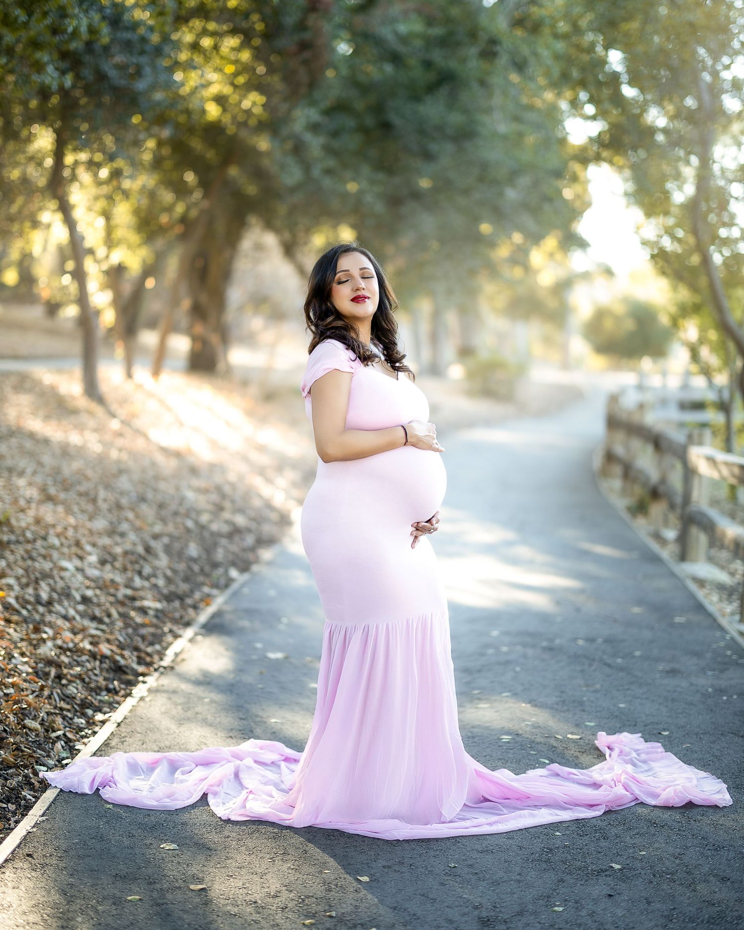 A person in a flowing pink maternity gown poses gracefully on a sunlit path surrounded by autumn leaves.