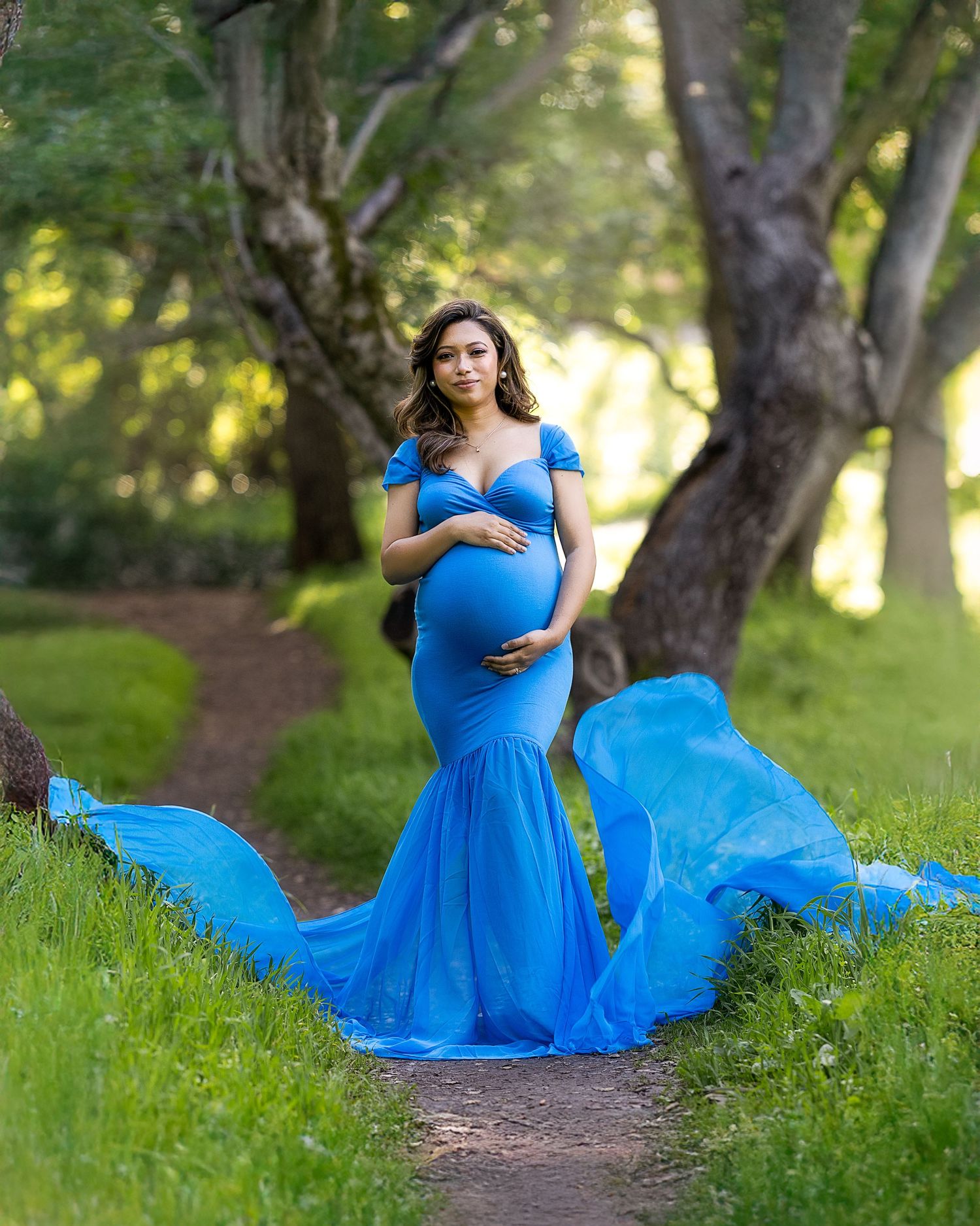 Graceful figure in flowing blue gown twirls through sunlit garden path surrounded by trees.