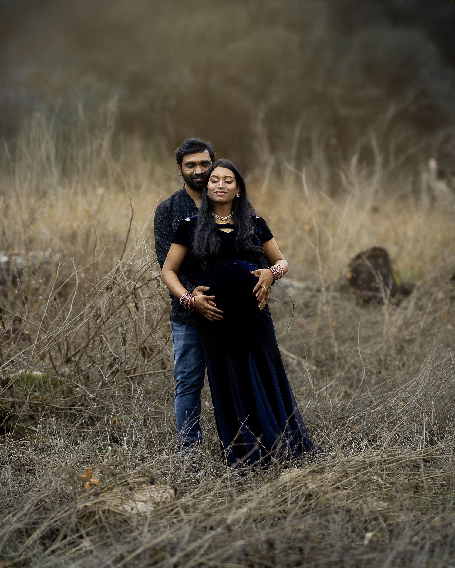A couple embraces in a romantic pose among tall dry grass in a moody outdoor setting.