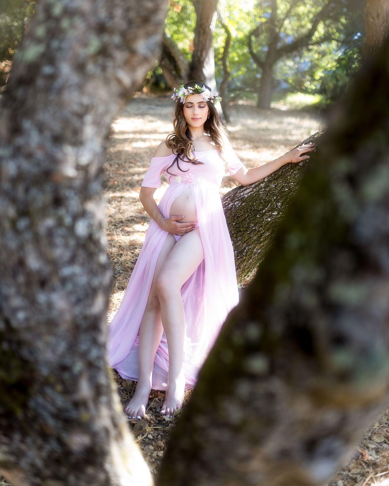 A person in a flowing pink dress poses gracefully against a tree trunk in dappled sunlight.