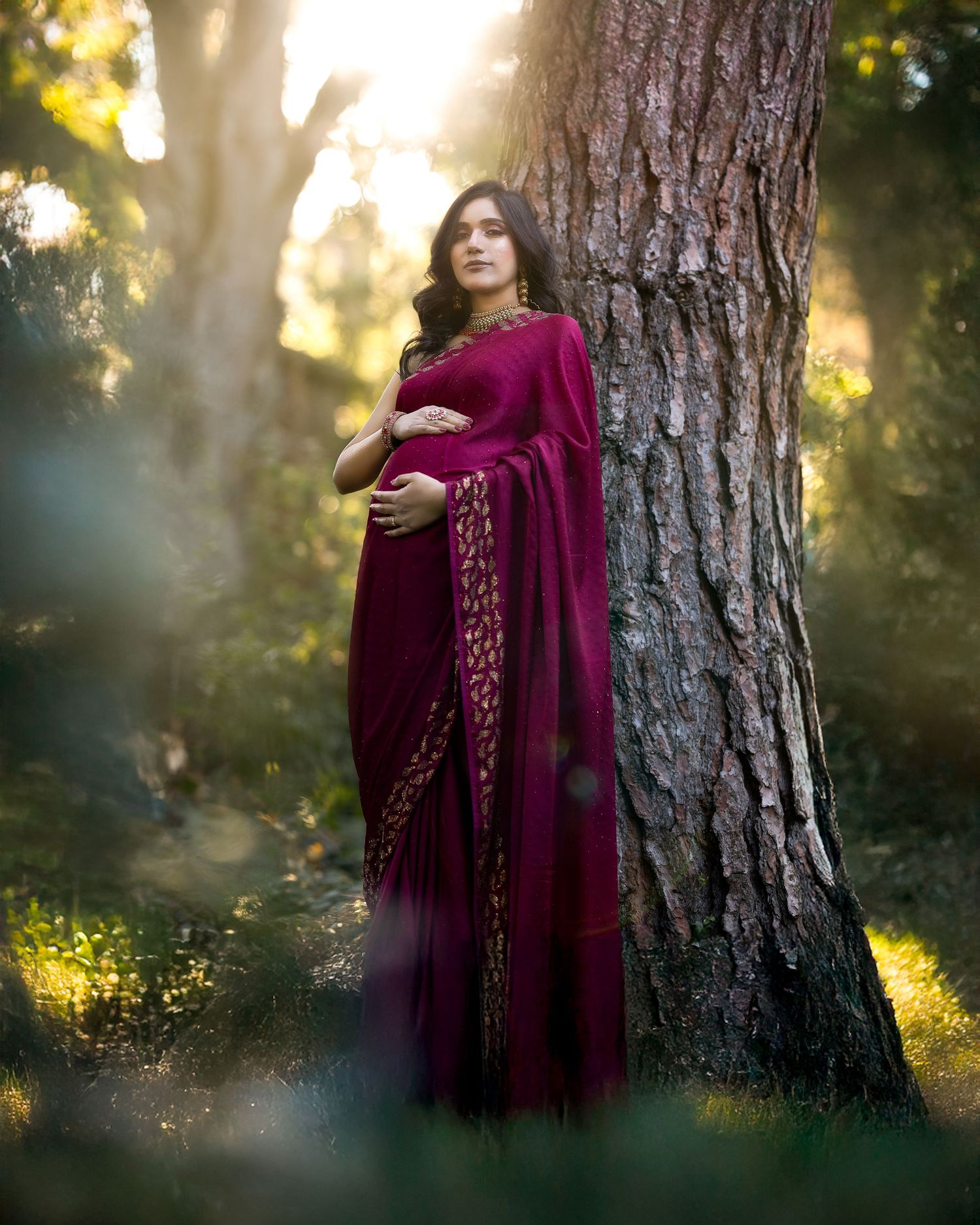 A person in a burgundy velvet dress poses against a tree trunk in dreamy golden sunlit forest setting.