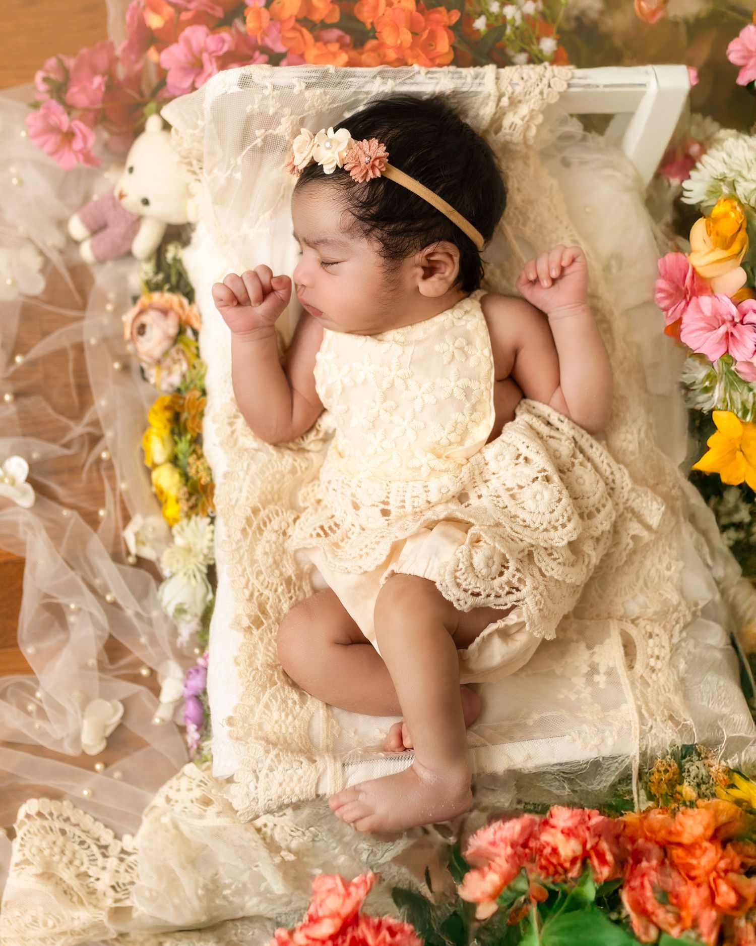 A sleeping baby in cream lace dress lies surrounded by colorful flowers on delicate tulle fabric.