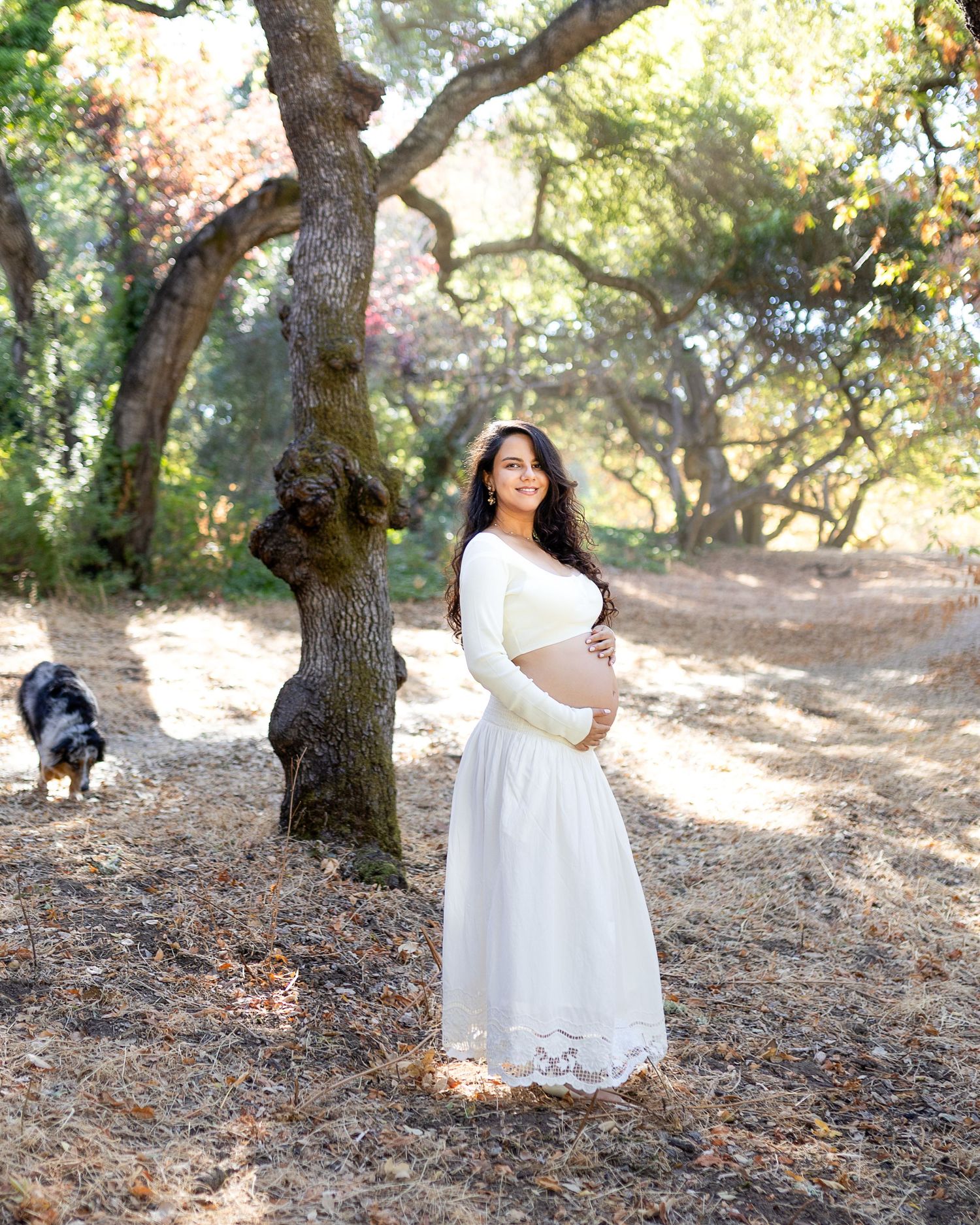 A white lace dress flows elegantly against a natural backdrop of trees and fallen autumn leaves in an outdoor setting.