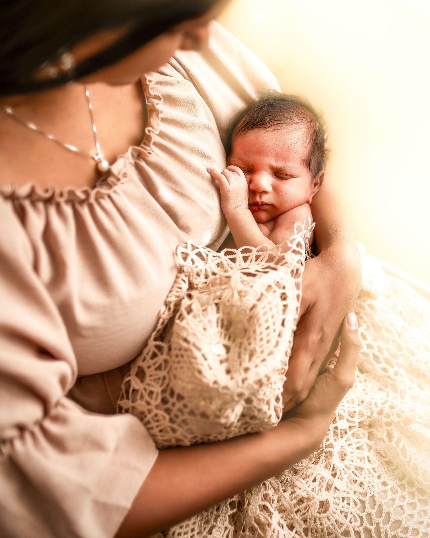 A newborn baby wrapped in delicate lace fabric rests against someone wearing a soft peach-colored dress.